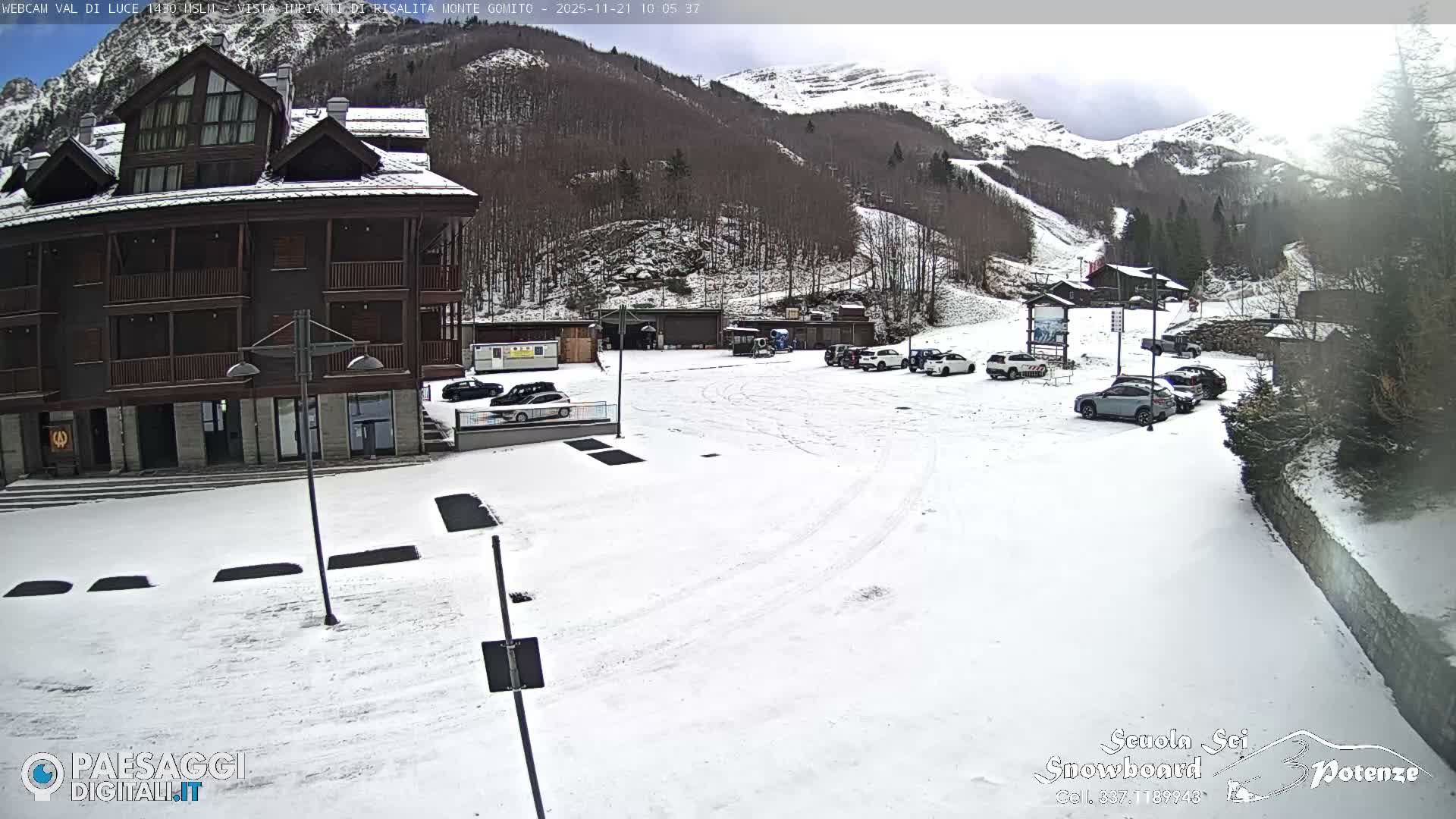 A snowy mountain resort on an overcast day shows a large lodge and a snow-covered parking lot with cars, backed by forested hillsides and rugged, snow-capped mountains.