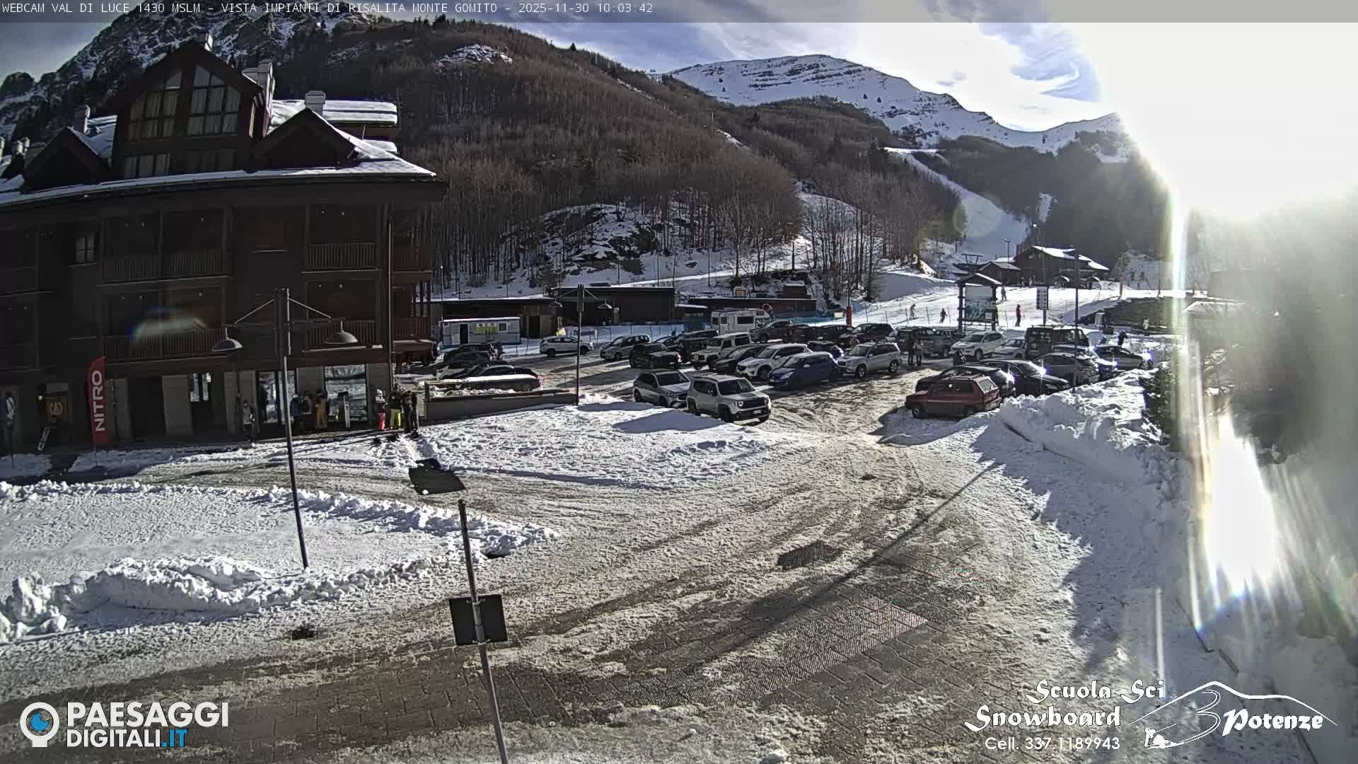 A sunny winter day illuminates a snow-covered mountain resort featuring a large building, a busy parking lot filled with vehicles, and distant ski slopes on the surrounding treed mountains under a clear sky.