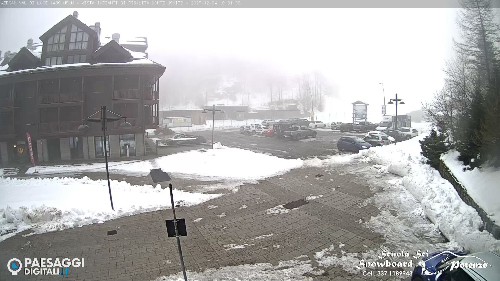 A multi-story building and a busy parking lot are visible amidst a snowy landscape on a heavily fogged winter day.
