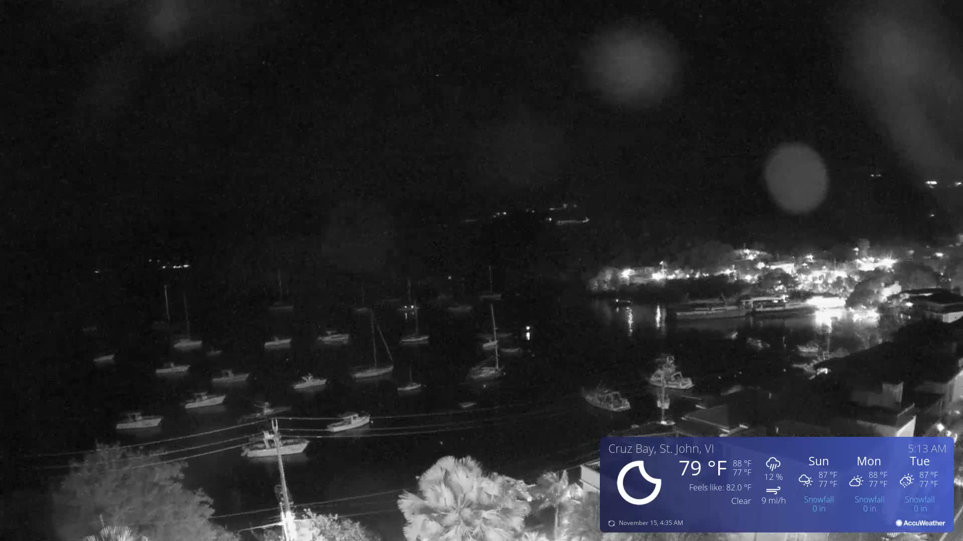 A clear night view overlooks a bay dotted with numerous anchored boats, leading to a brightly lit townscape along the distant shore and hillside.