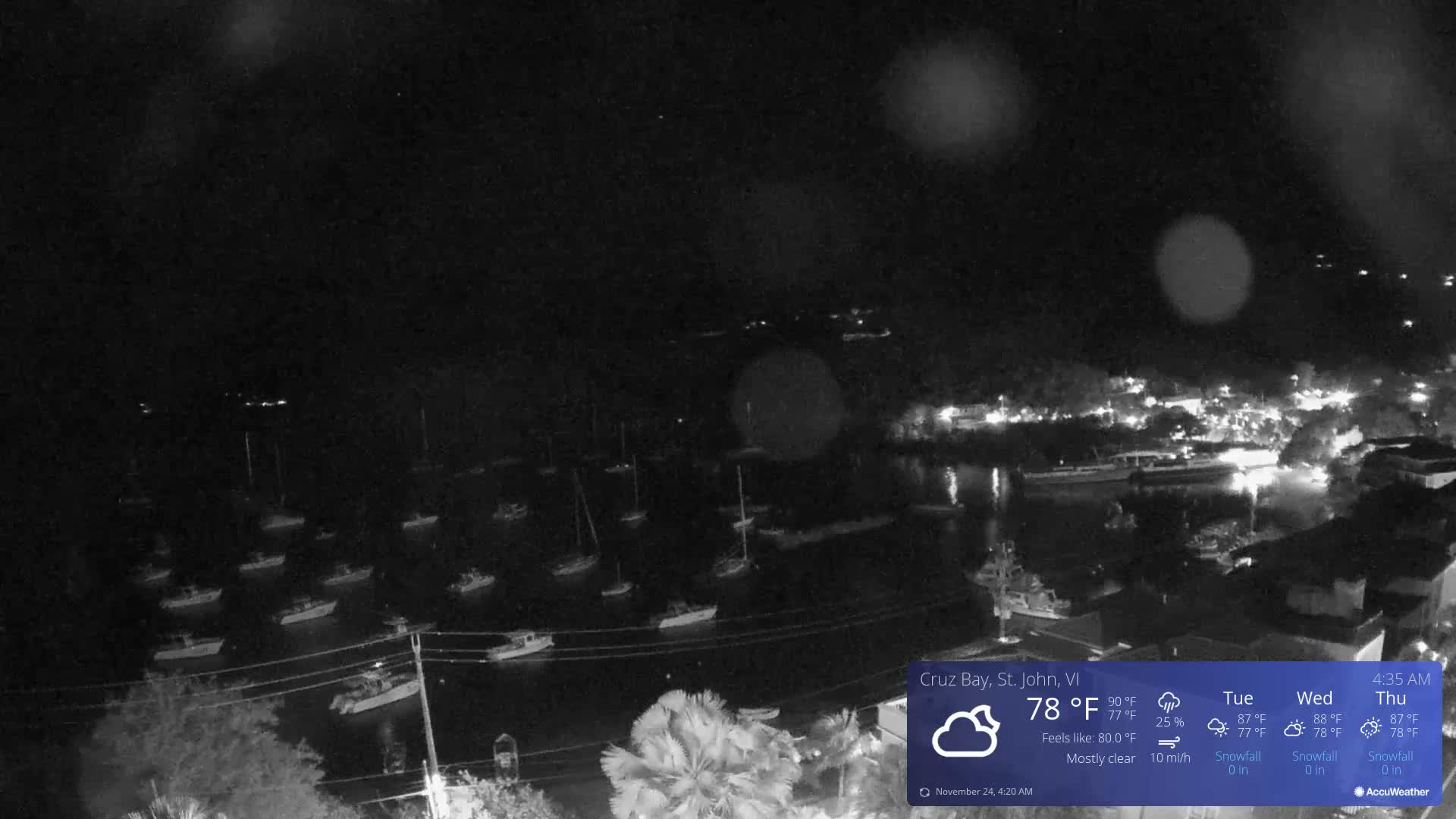 A dark, rainy night view captures a bay dotted with numerous moored boats, while the lights of a town illuminate the distant shore.