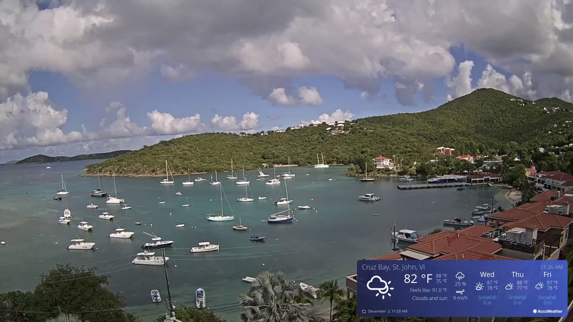 A wide view of a vibrant bay filled with numerous boats and sailboats, surrounded by lush green hills topped with scattered buildings and houses under a partly cloudy sky.