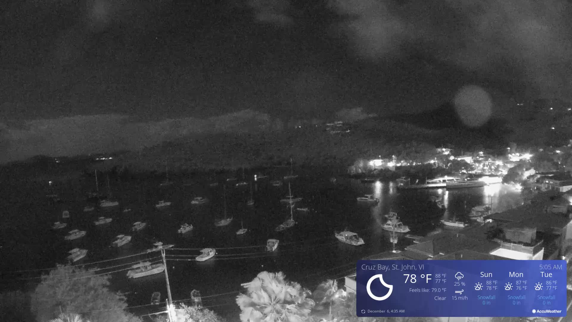 A grayscale nighttime image presents a wide view of a bay densely populated with various boats at anchor, with a softly lit coastline featuring buildings to the right and obscured hills in the distance, all beneath a dark, hazy sky.