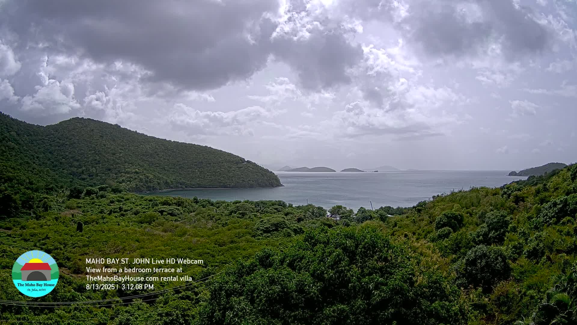 A tranquil bay is seen from a high vantage point, under a cloudy sky, with lush green hills and islands in the distance.
