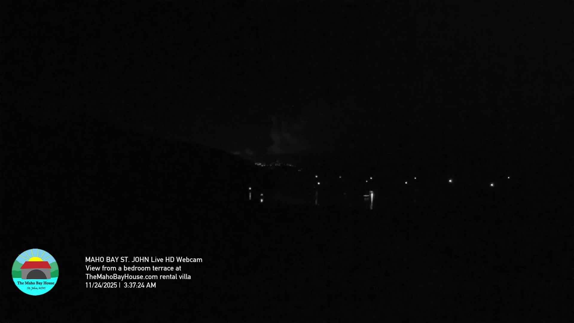 A dark, cloudy night scene reveals faint outlines of distant hills dotted with lights, overlooking a body of water featuring several illuminated points.