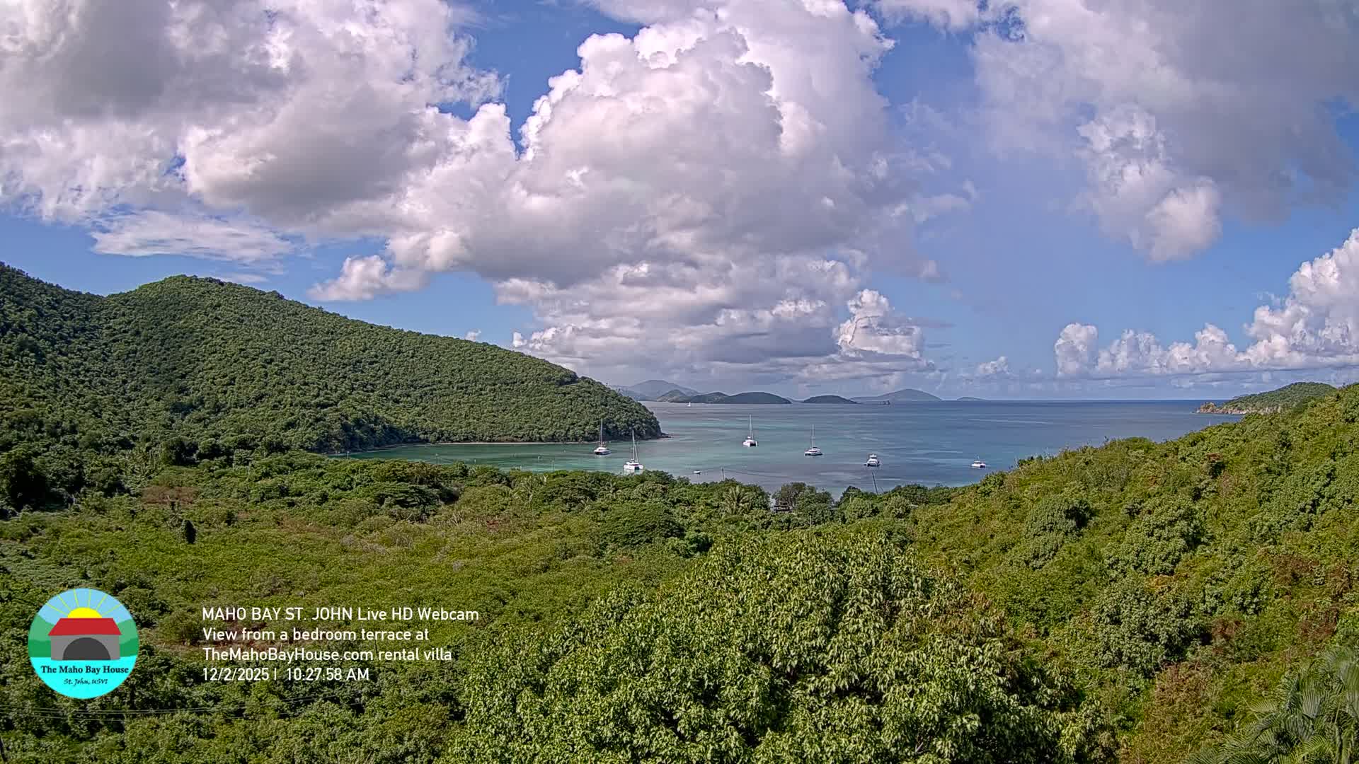 The image shows a sunny day with scattered white clouds over a tranquil tropical bay dotted with several anchored sailboats, all framed by lush green, forested hills.