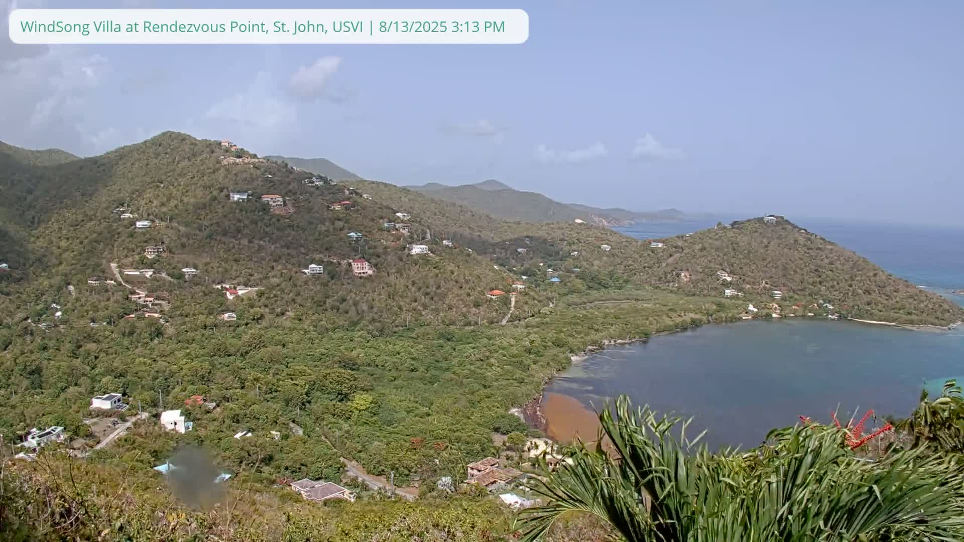 A high-angle, long shot shows a clear, sunny day overlooking a tropical bay nestled between lush green hills dotted with houses, with the ocean visible in the background.