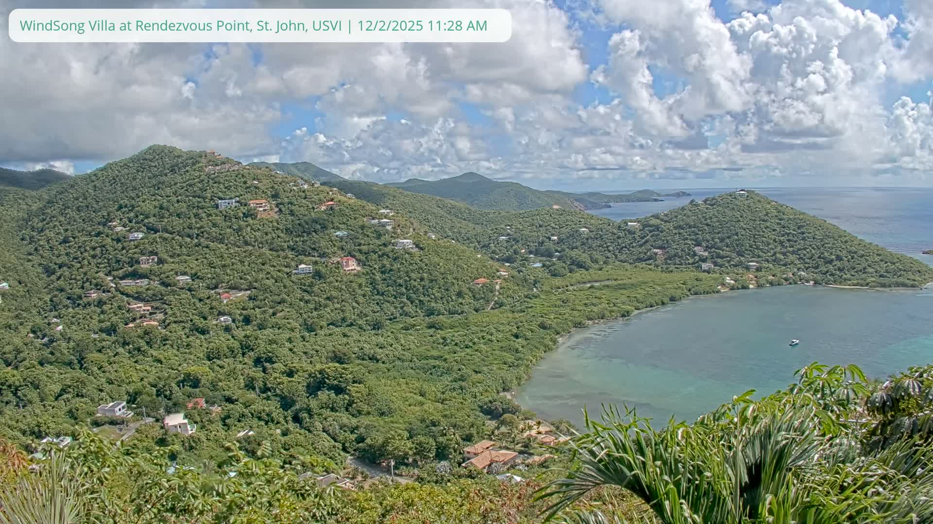 A vibrant landscape shot under a partly cloudy sky showcases densely forested hills studded with numerous houses, curving around a tranquil turquoise bay where a single boat floats.