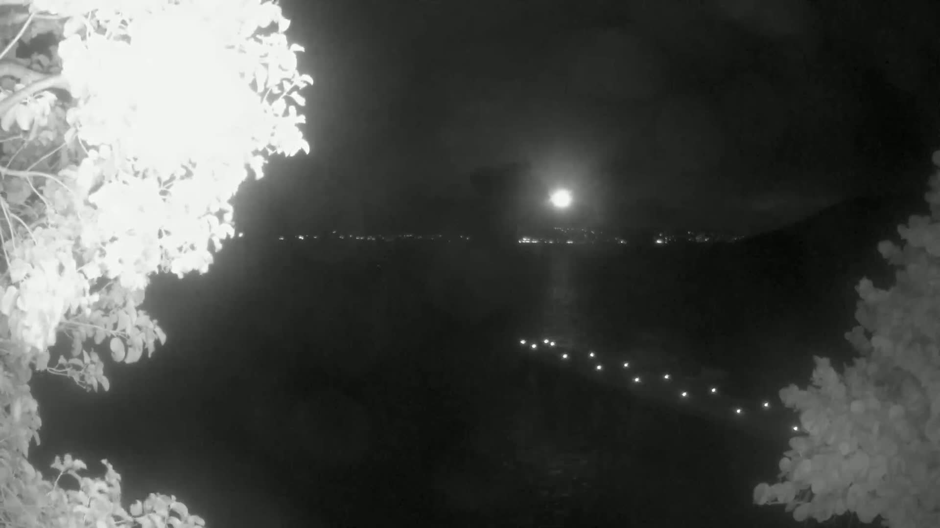 A nighttime view shows a body of water with lights on a pier in the foreground and a bright moon over a distant shoreline.