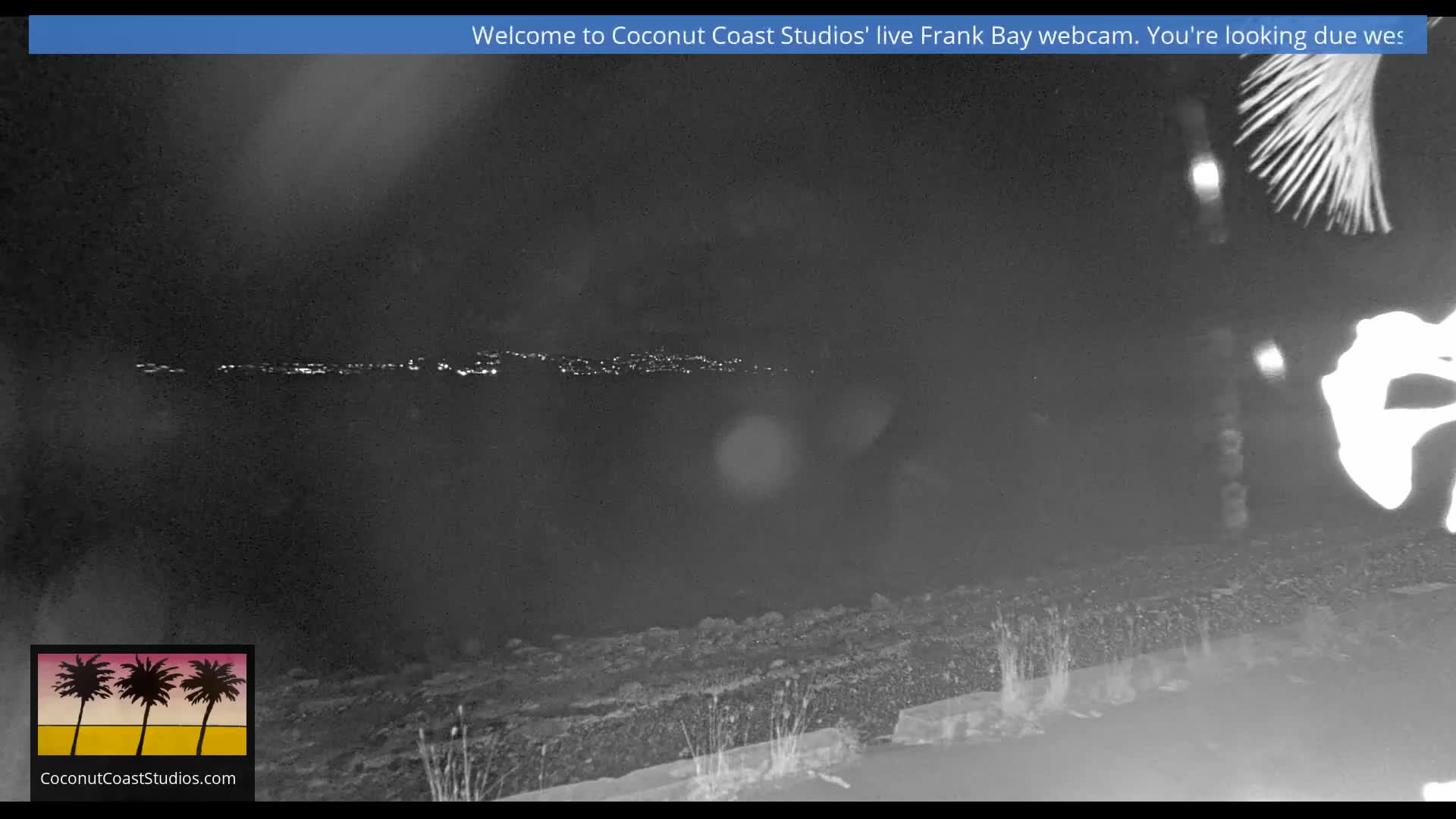 A clear night view captures a dark body of water reflecting the distant twinkling lights of a far-off shoreline, with a dark, textured foreground and bright, abstract reflections or water spray on the lower right.