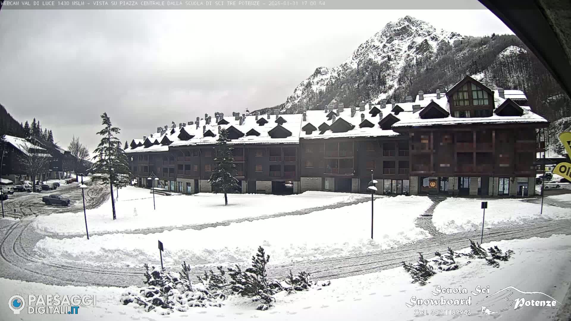 A snow-covered plaza sits in front of a multi-story building, with a mountain range in the background under an overcast sky.