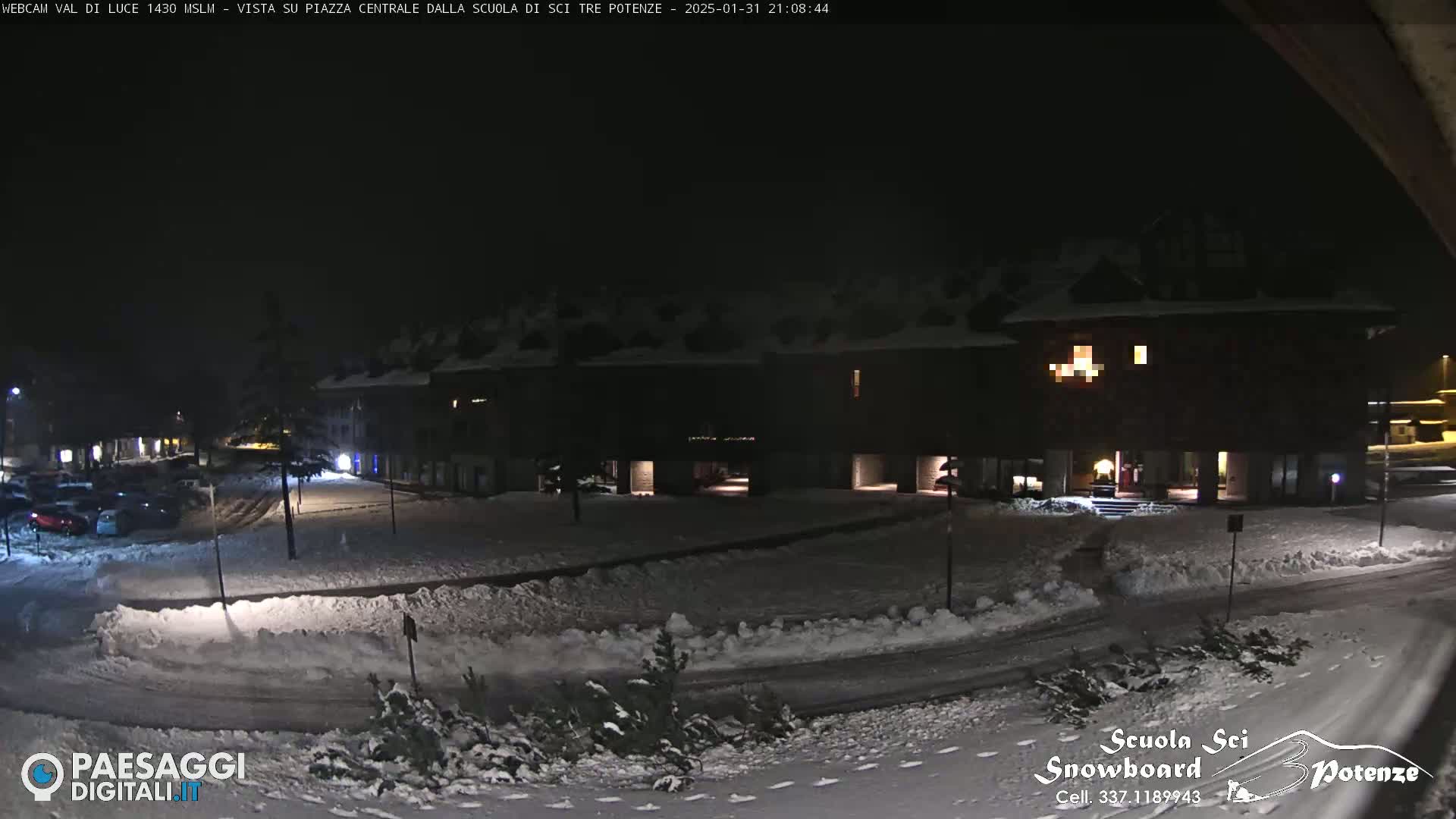 A snow-covered plaza at night, illuminated by building lights, is observed from a slightly elevated vantage point.