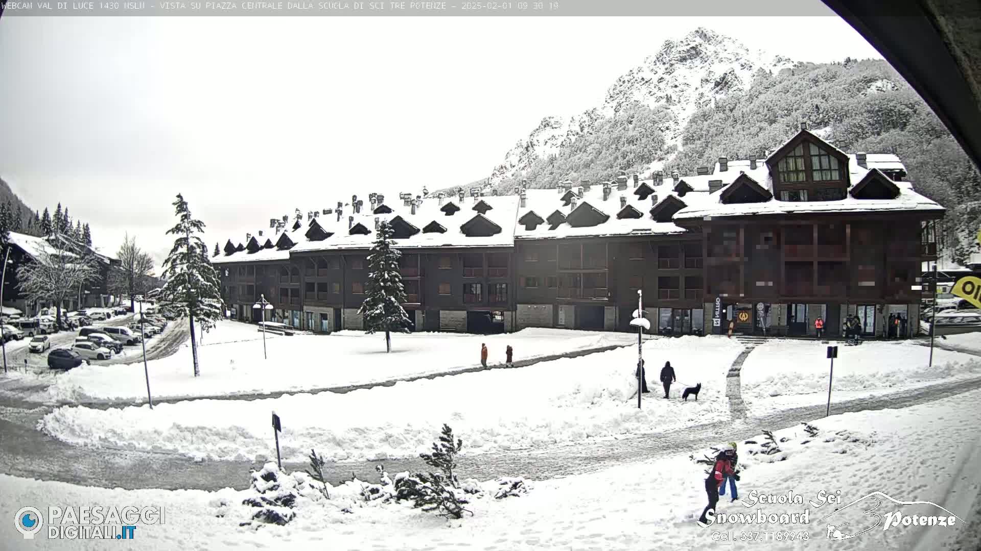 A snow-covered town square sits in front of a large, multi-story building, with snow-capped mountains in the background under an overcast sky.