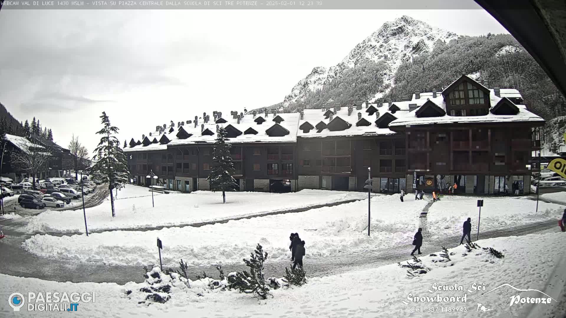 A snow-covered plaza with a multi-story building and several people walking, situated in front of a mountain range under an overcast sky.