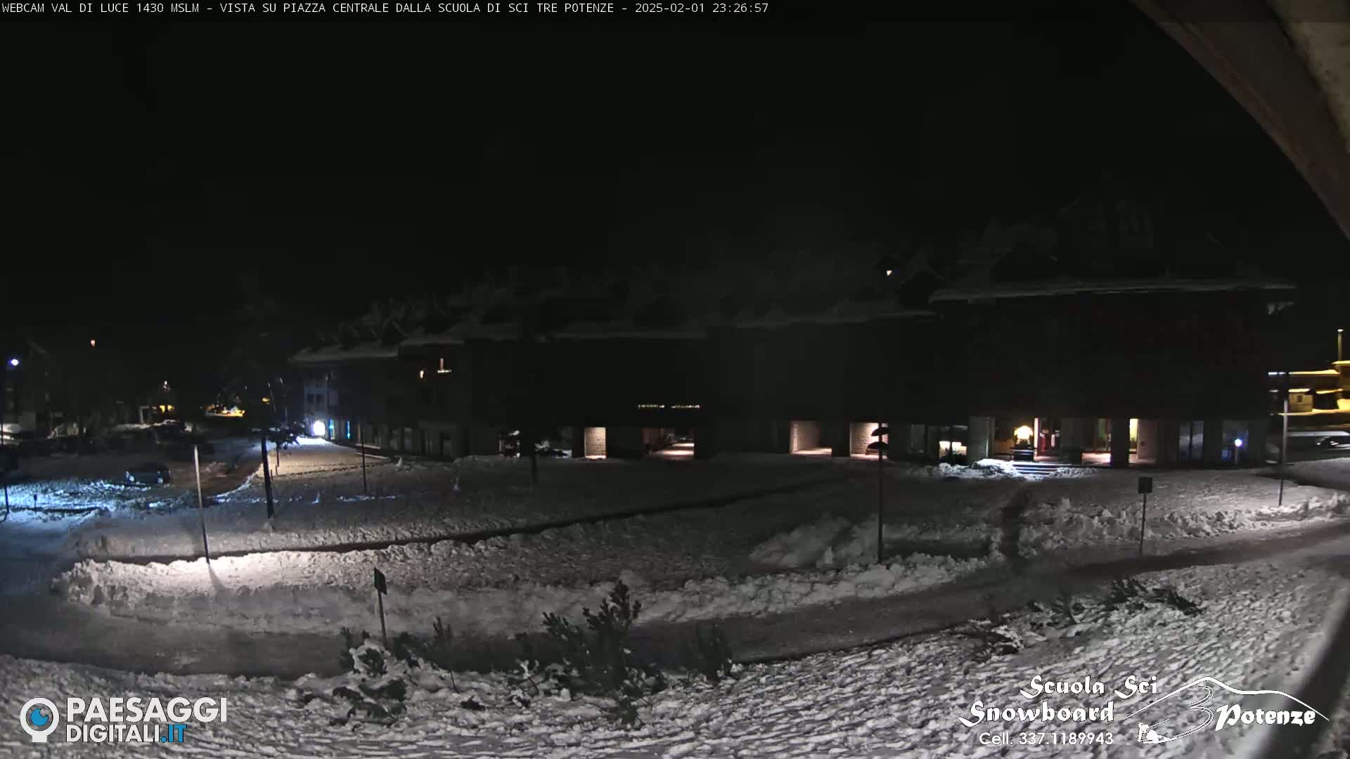 A snow-covered plaza at night, illuminated by building lights, is observed near a dark building.