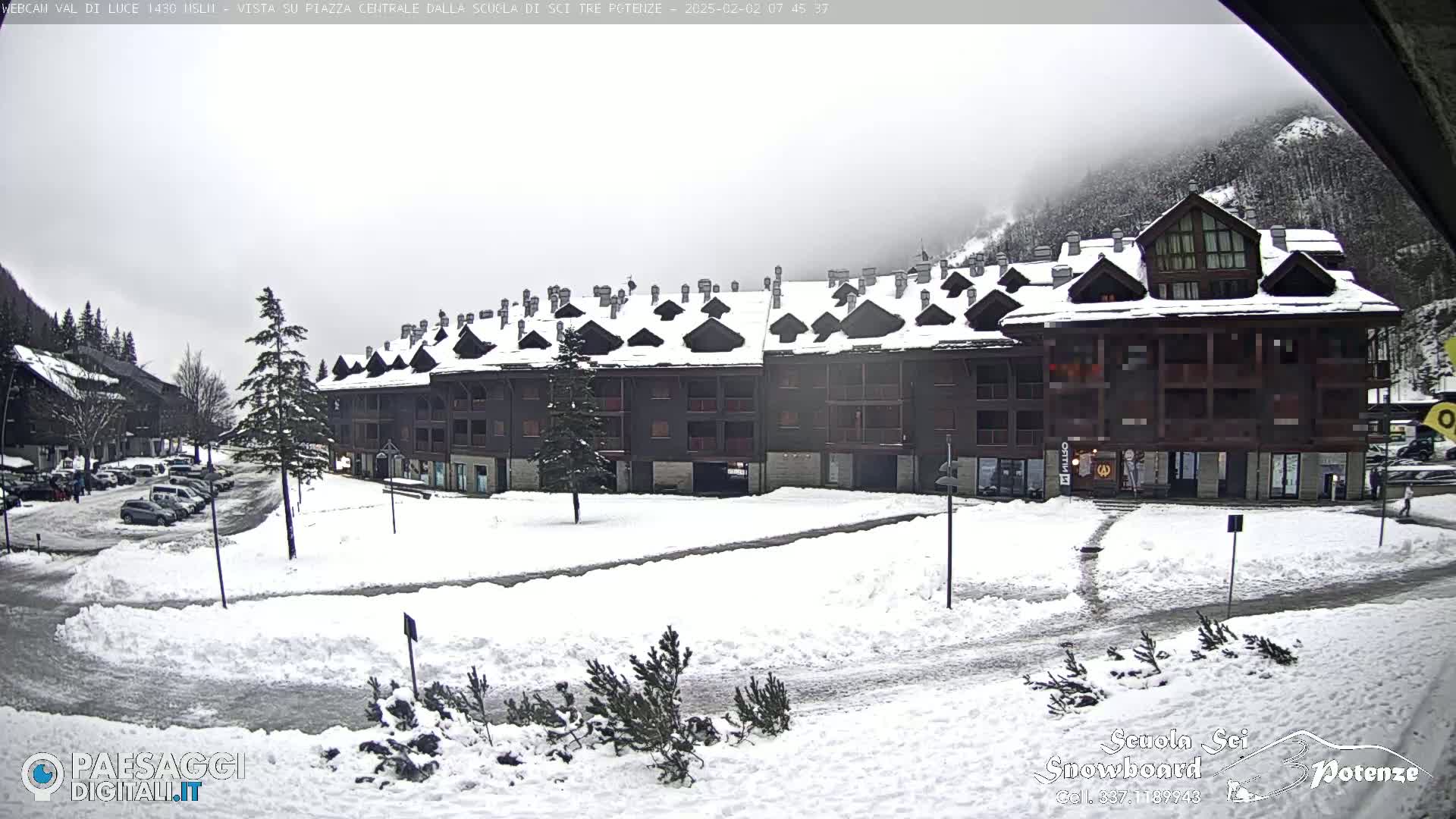 A snow-covered parking lot and plaza are in front of a multi-story building in a mountainous region under an overcast sky.