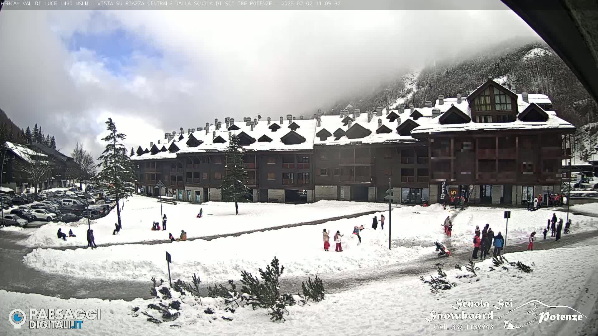 A snow-covered plaza with a large building and several people playing in the snow, under a mostly cloudy sky.