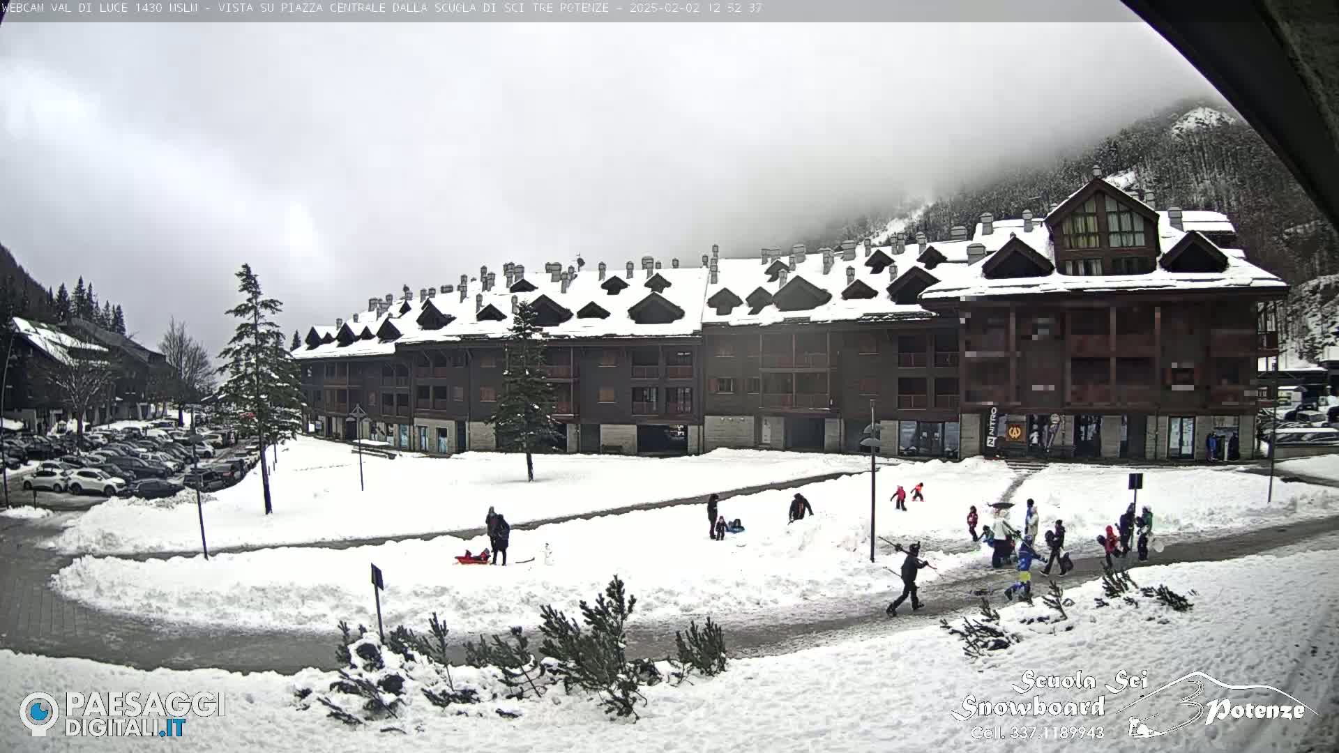 A snowy plaza with a multi-story building and several people, some sledding, in overcast weather.