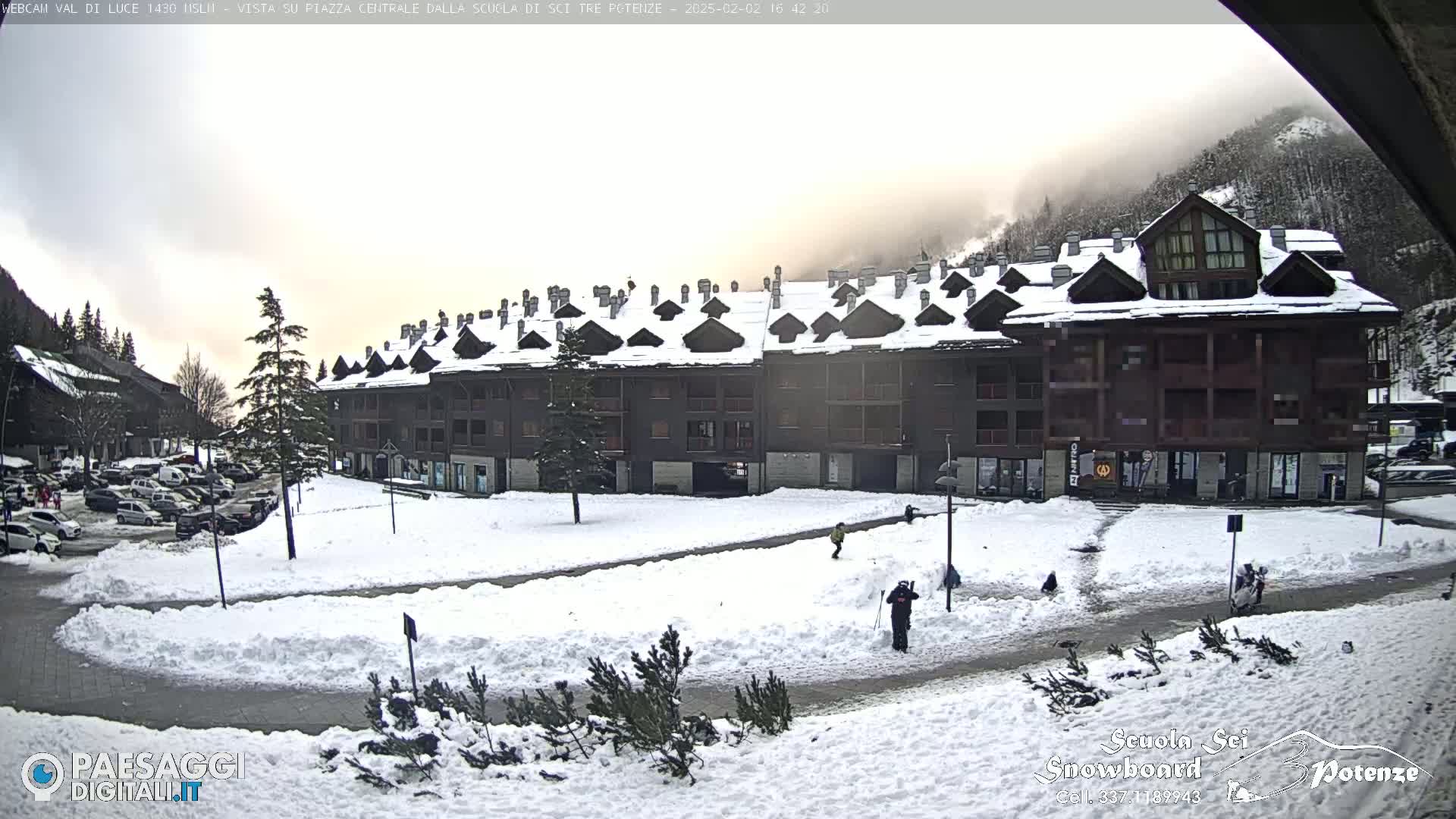 A snow-covered plaza in front of a multi-story building with a mountain backdrop under an overcast sky.