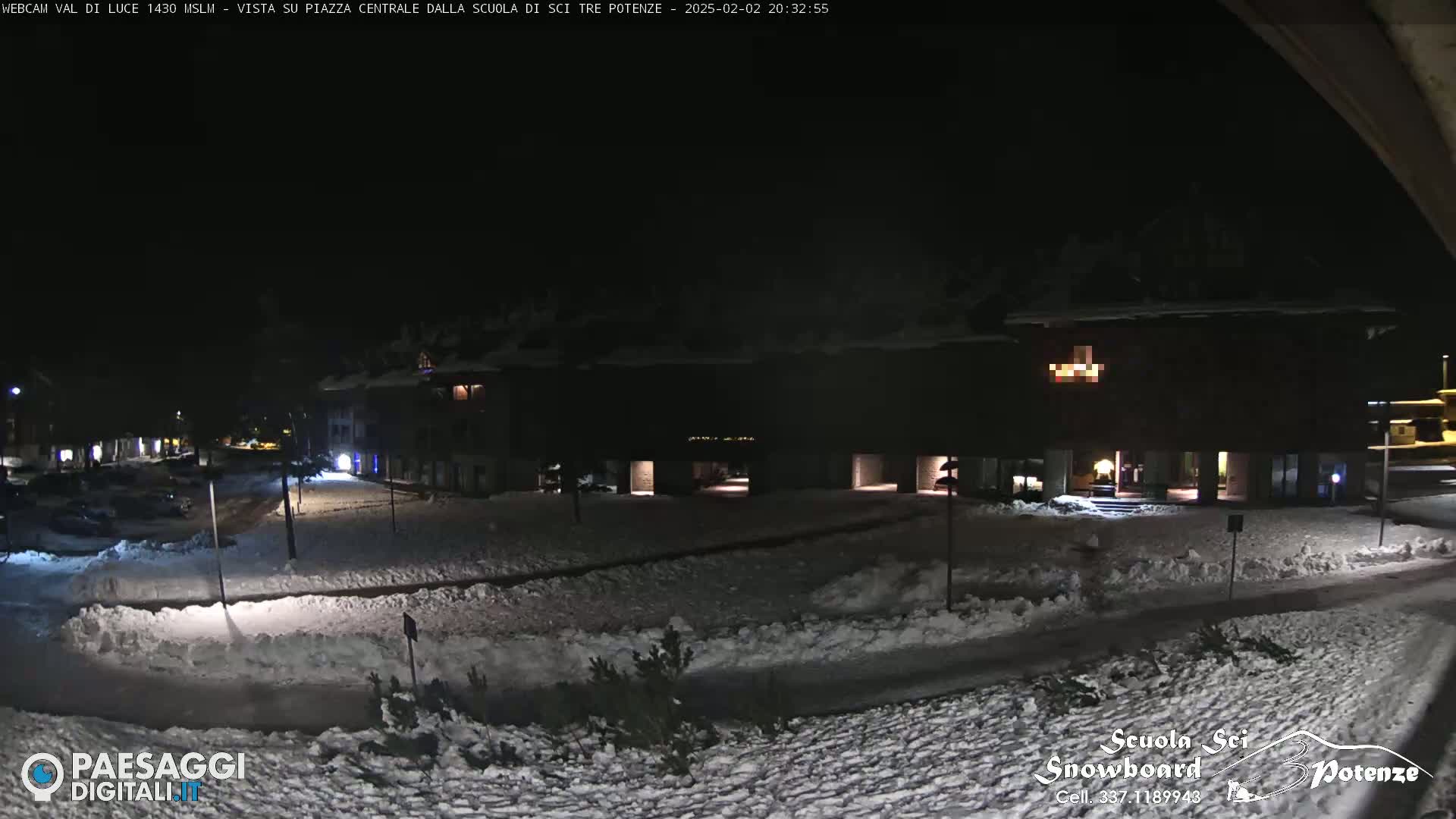 A snow-covered nighttime scene shows a town square with buildings and cars parked, illuminated by streetlights.