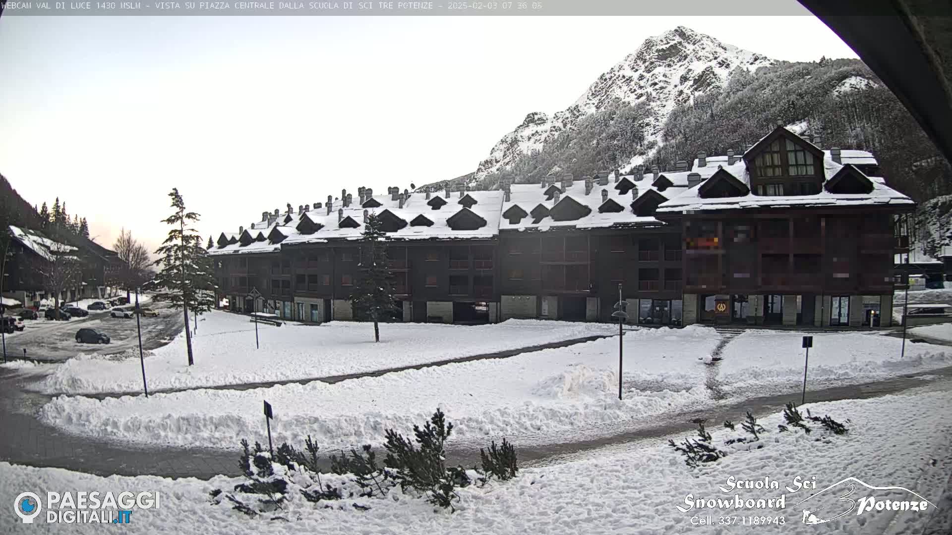 A snow-covered town square sits in front of a large, multi-unit building with a mountain in the background under a cloudy sky.