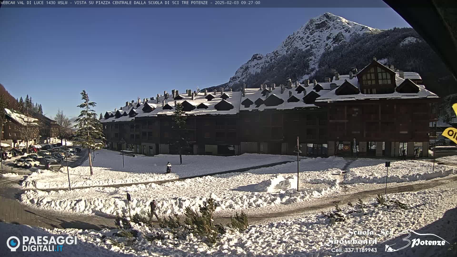 A snow-covered town square is situated in front of a large building, with a snow-capped mountain in the background under a clear blue sky.