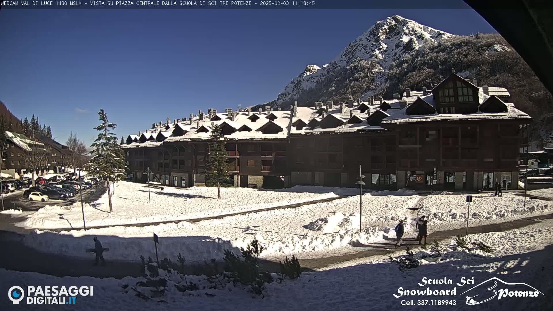 A snow-covered plaza sits in front of a large building complex, with a snow-capped mountain in the background under a clear blue sky.