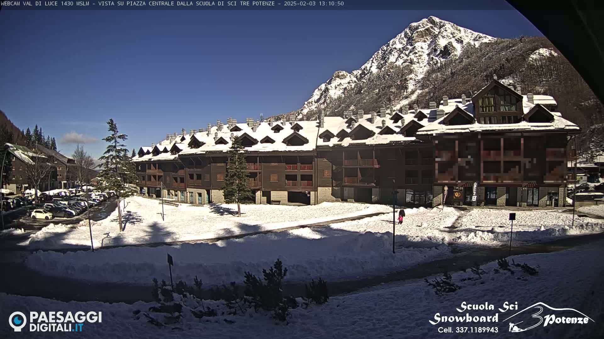 A snow-covered plaza with a multi-story building sits before a mountain range under a sunny sky.