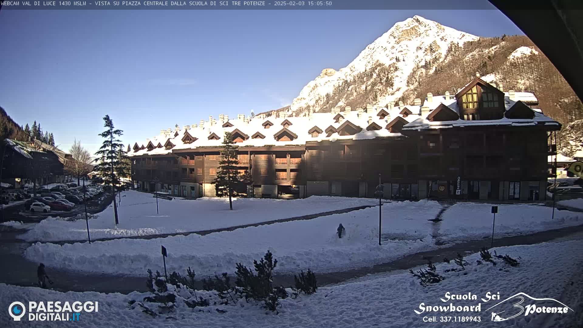 A snow-covered plaza with a multi-story building and a mountain in the background under a sunny sky.