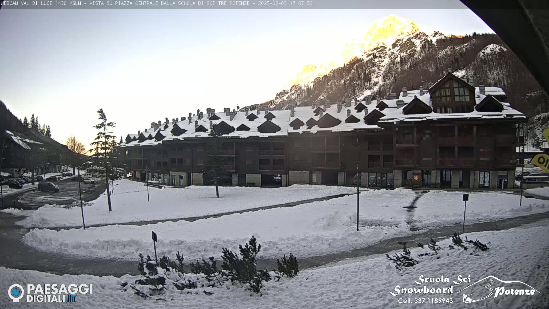A snow-covered plaza sits in front of a multi-story brown building, with mountains in the background under a mostly clear sky, illuminated by the setting sun.