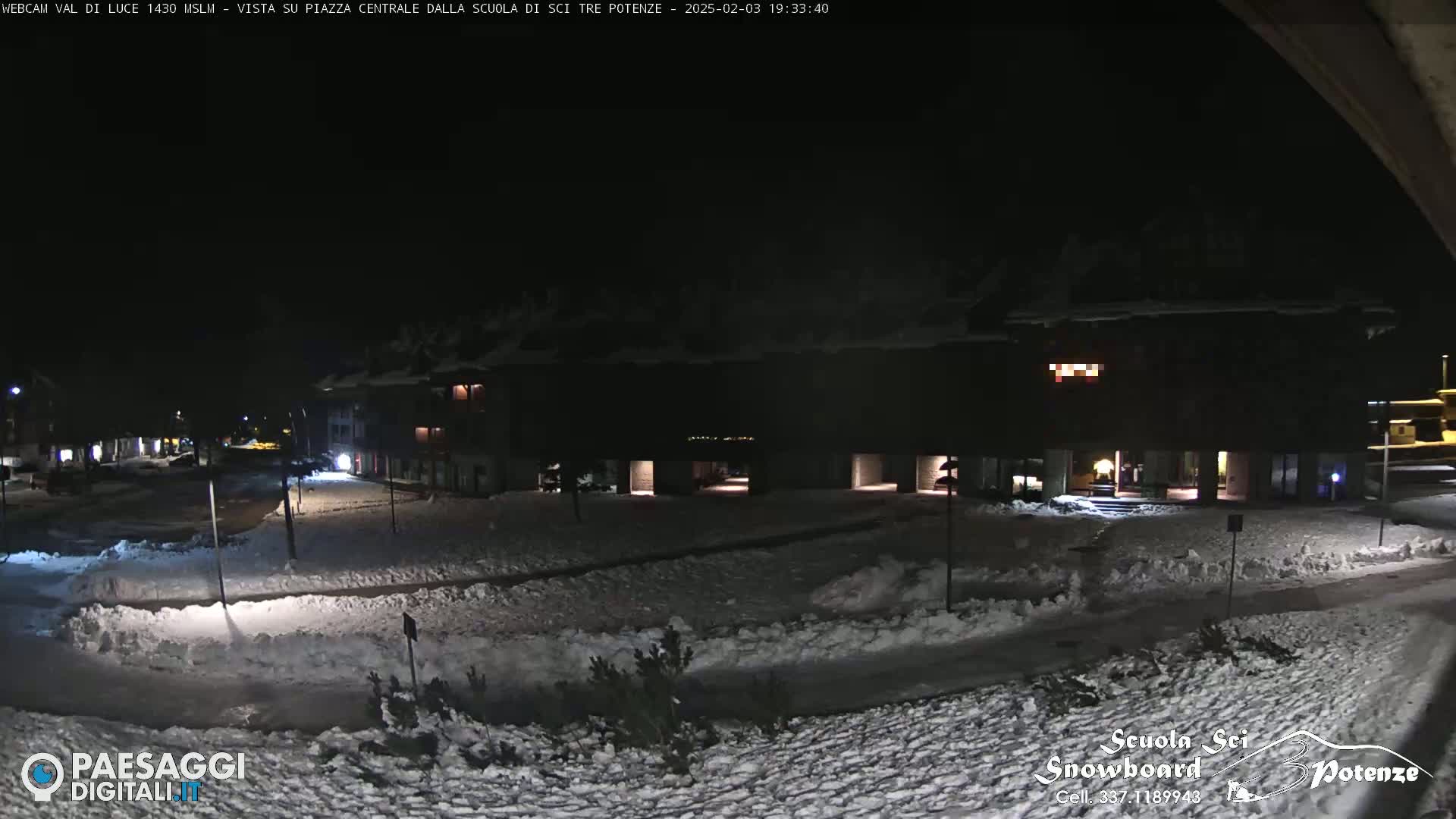 A snow-covered town square at night, illuminated by streetlights, with buildings lining the perimeter.