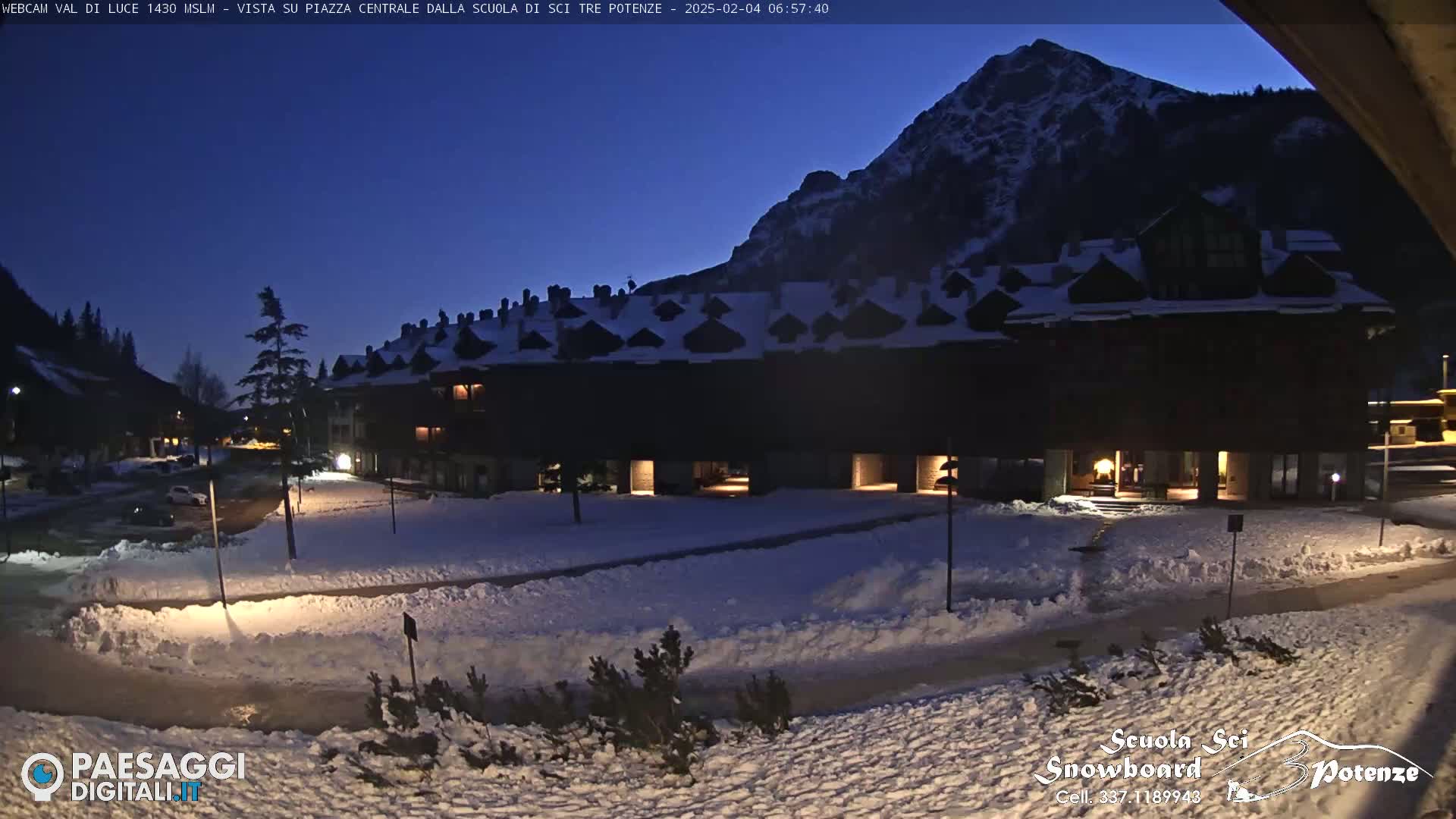 A snow-covered plaza at twilight, with a large building illuminated against a backdrop of a dark mountain.