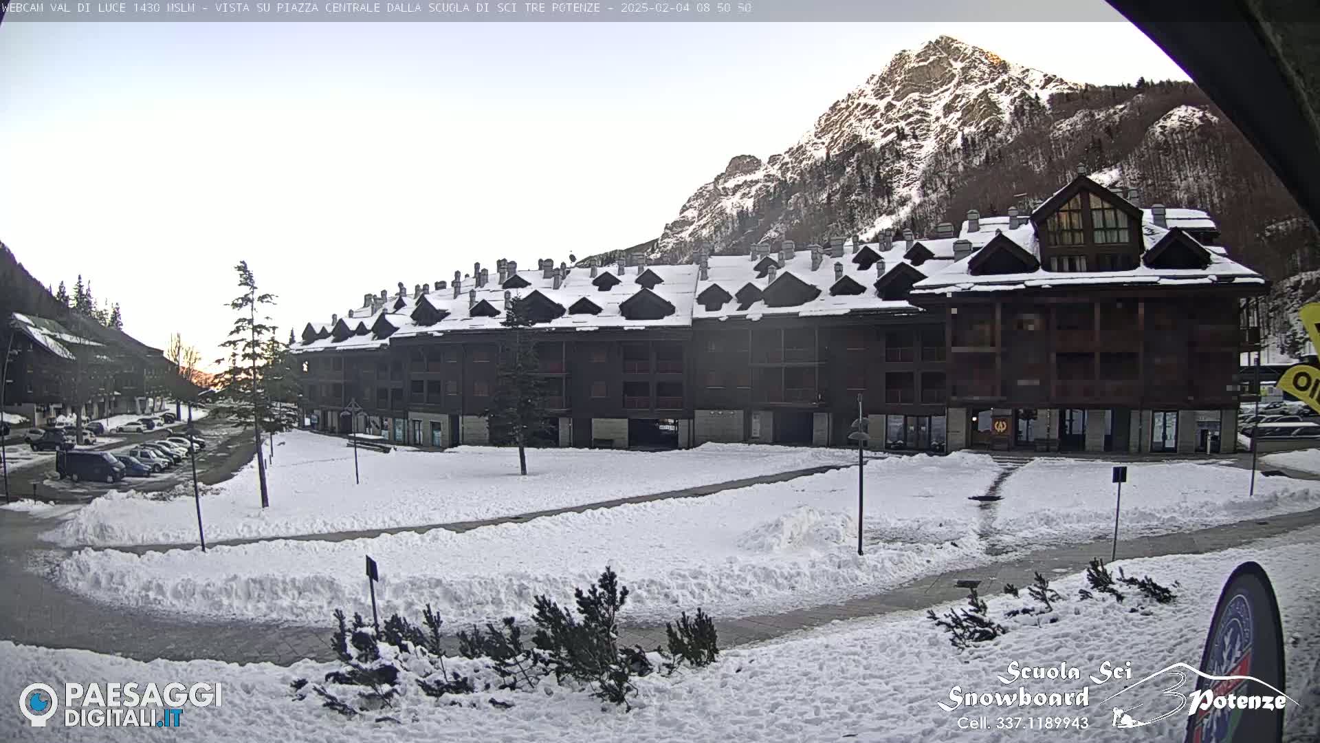 A snow-covered plaza sits in front of a multi-story brown building, with mountains in the background under a mostly clear sky.