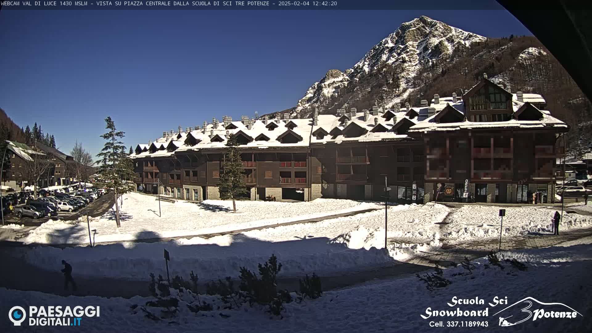 A snow-covered plaza in front of a large, multi-story building with a mountain in the background under a clear, sunny sky.