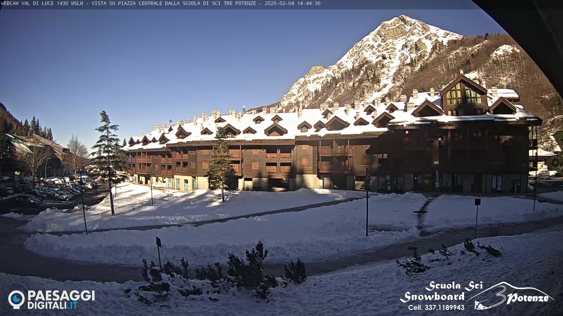 A snow-covered plaza sits in front of a large, multi-story building in the shadow of a sunlit mountain on a clear, sunny day.