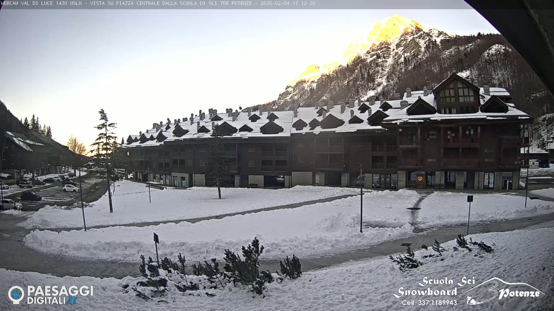A snow-covered plaza with a multi-story building in the background, mountains in the distance, and a setting sun.