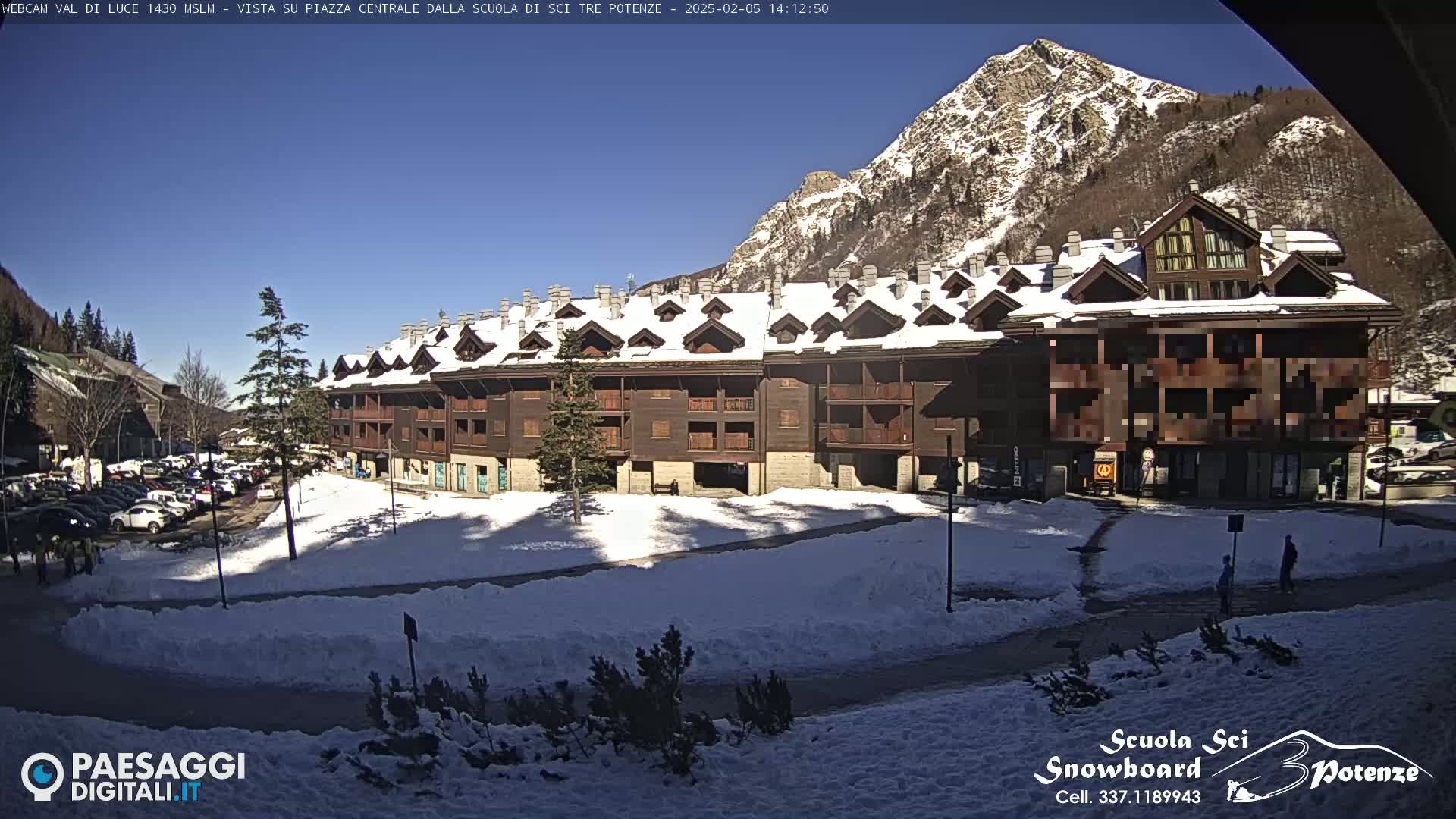 A long, multi-story brown building with snow-covered roofs sits in a snowy plaza, backed by a snow-dusted mountain range under a clear blue sky.
