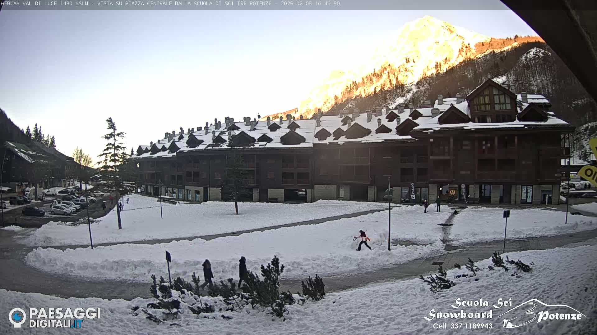 A snow-covered plaza with a large brown building in front of a sunlit mountain.
