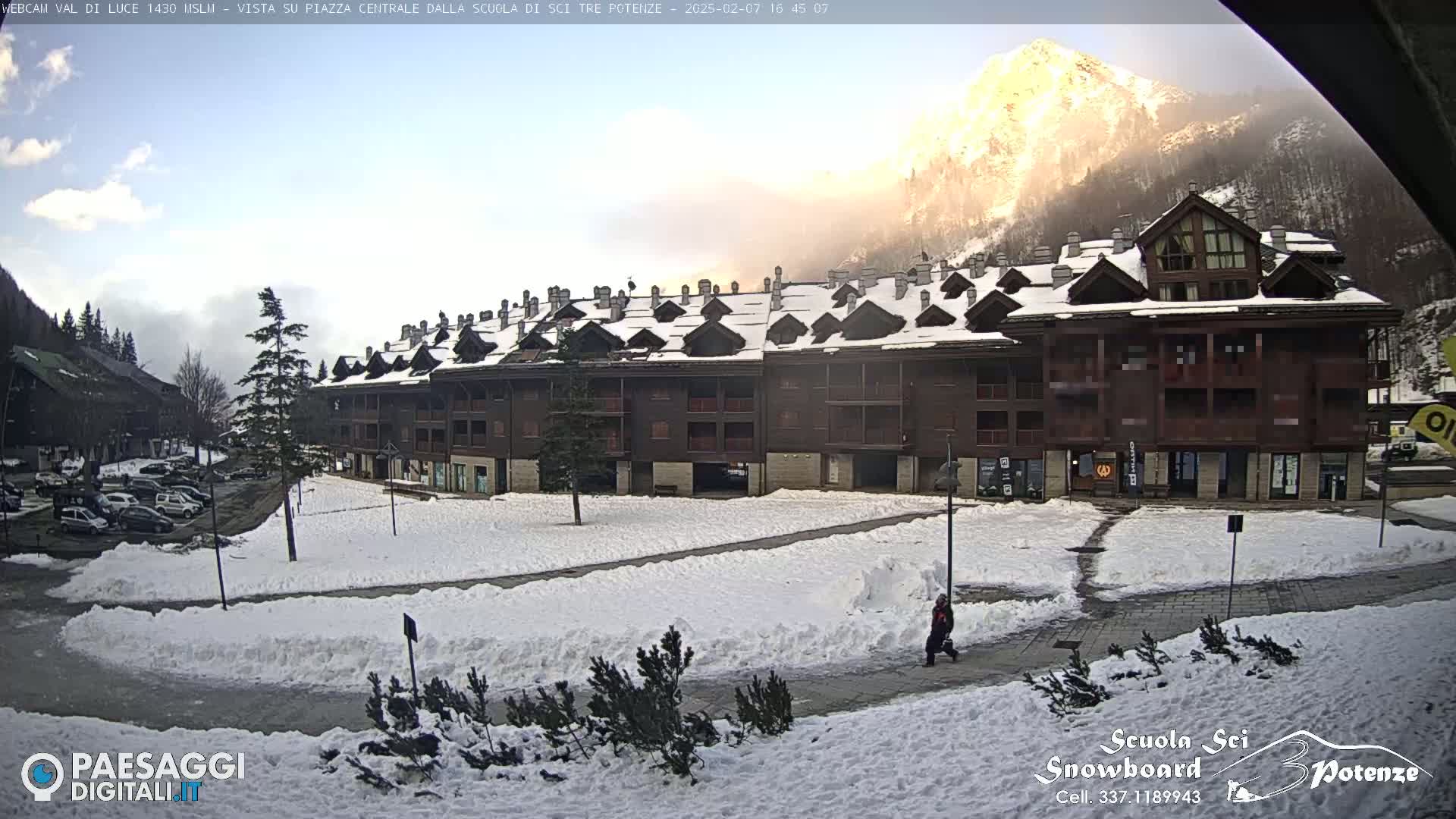 A snow-covered plaza is seen in front of a multi-story brown building, with a sunlit mountain in the background.