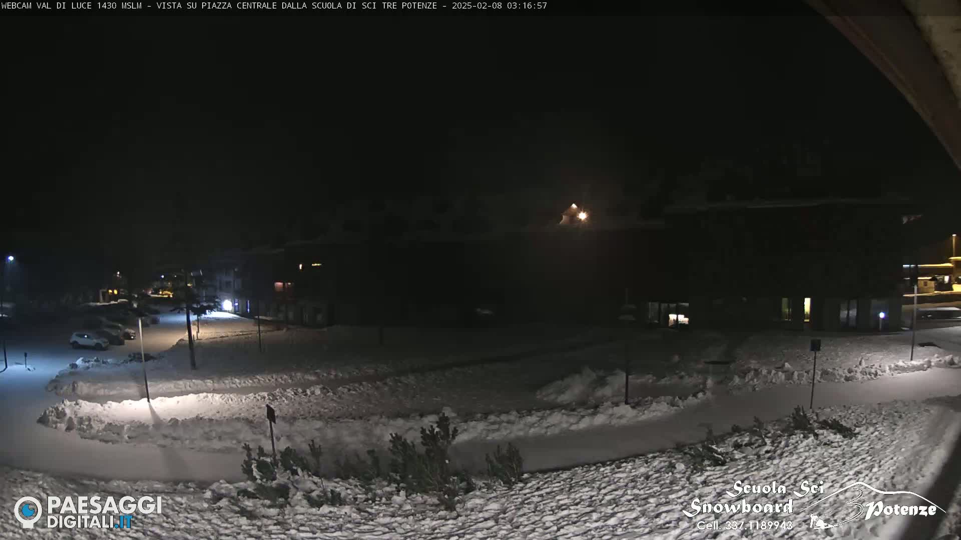 A snow-covered parking lot and plaza are dimly lit at night, with a few cars visible and buildings in the background.