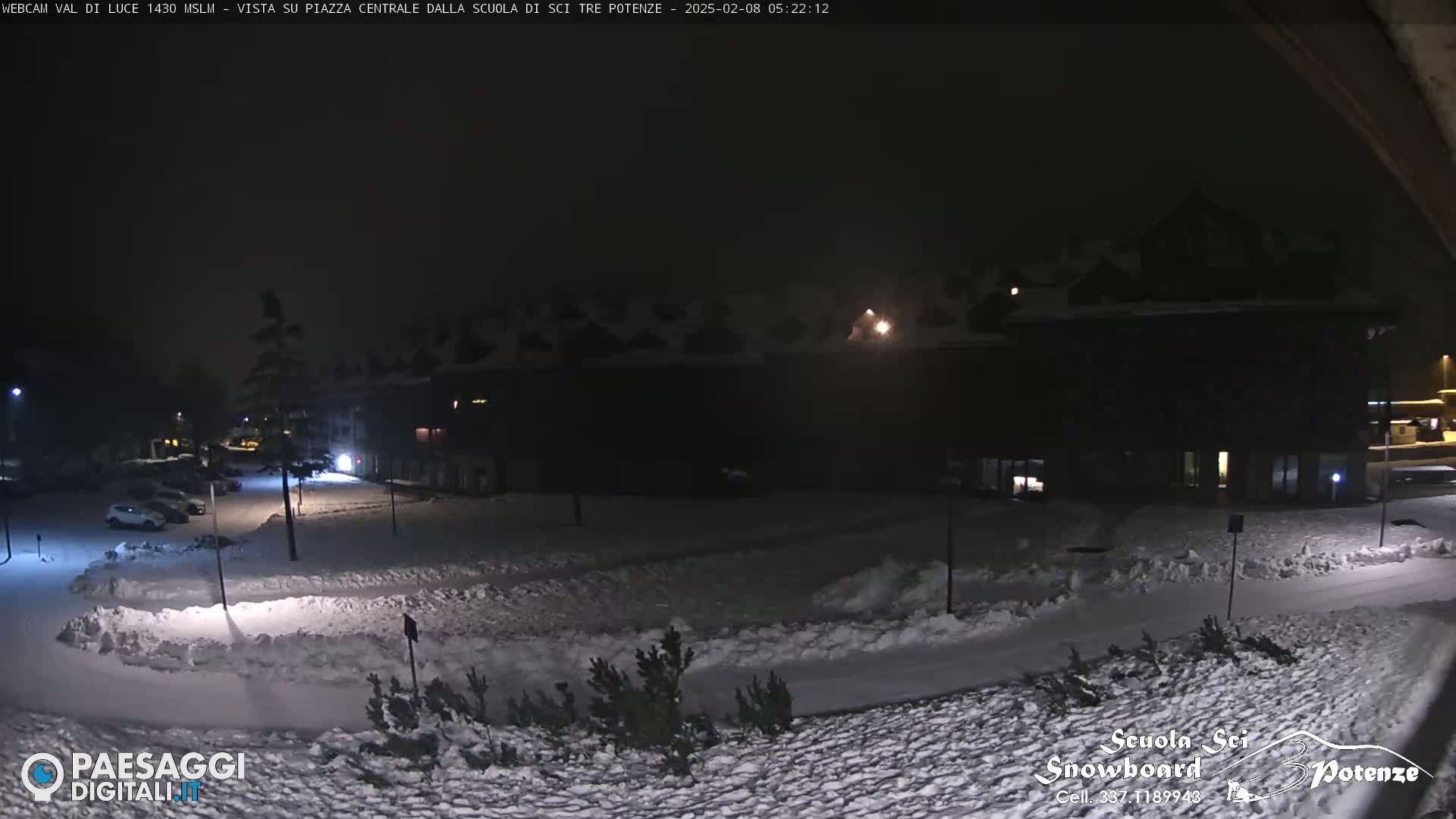 A snow-covered nighttime scene shows a parking lot with cars, small trees, and a large building, illuminated by artificial light.