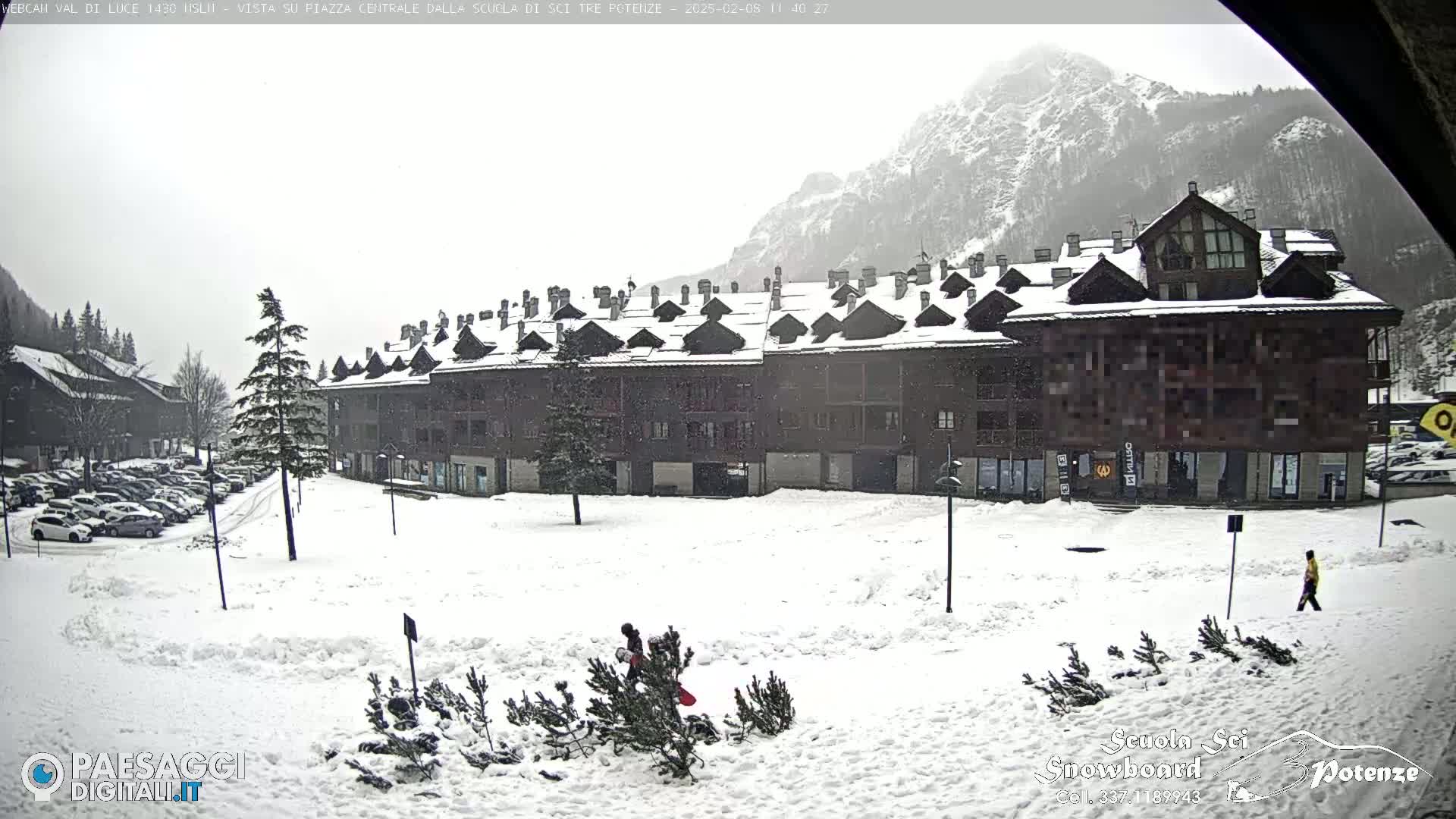 A large, snow-covered building with numerous windows and pitched roofs sits in a snowy square, with a mountain visible in the background under a heavily overcast sky.