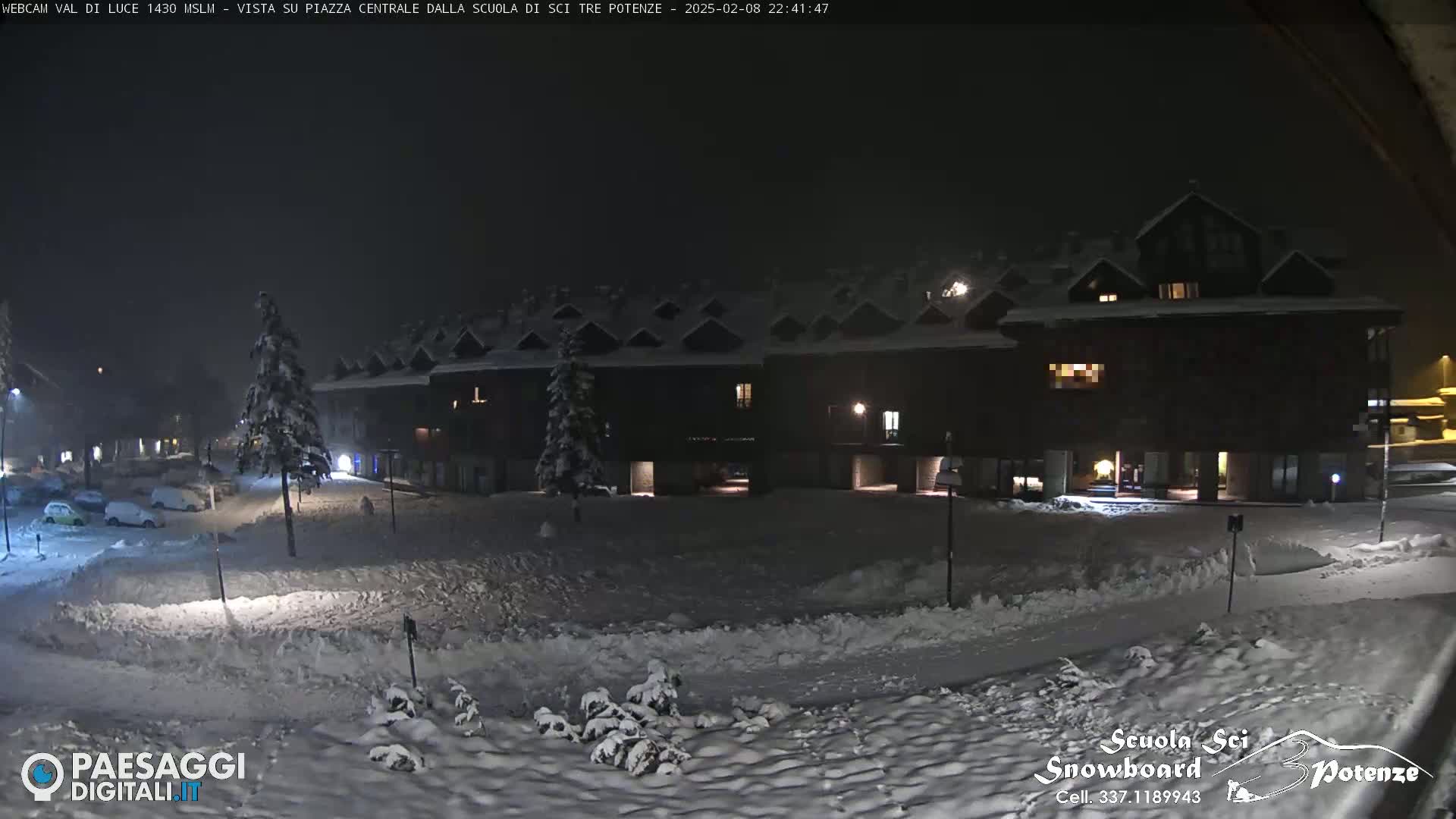 A snow-covered plaza at night, illuminated by streetlights, is situated in front of a dark, snow-dusted building complex.