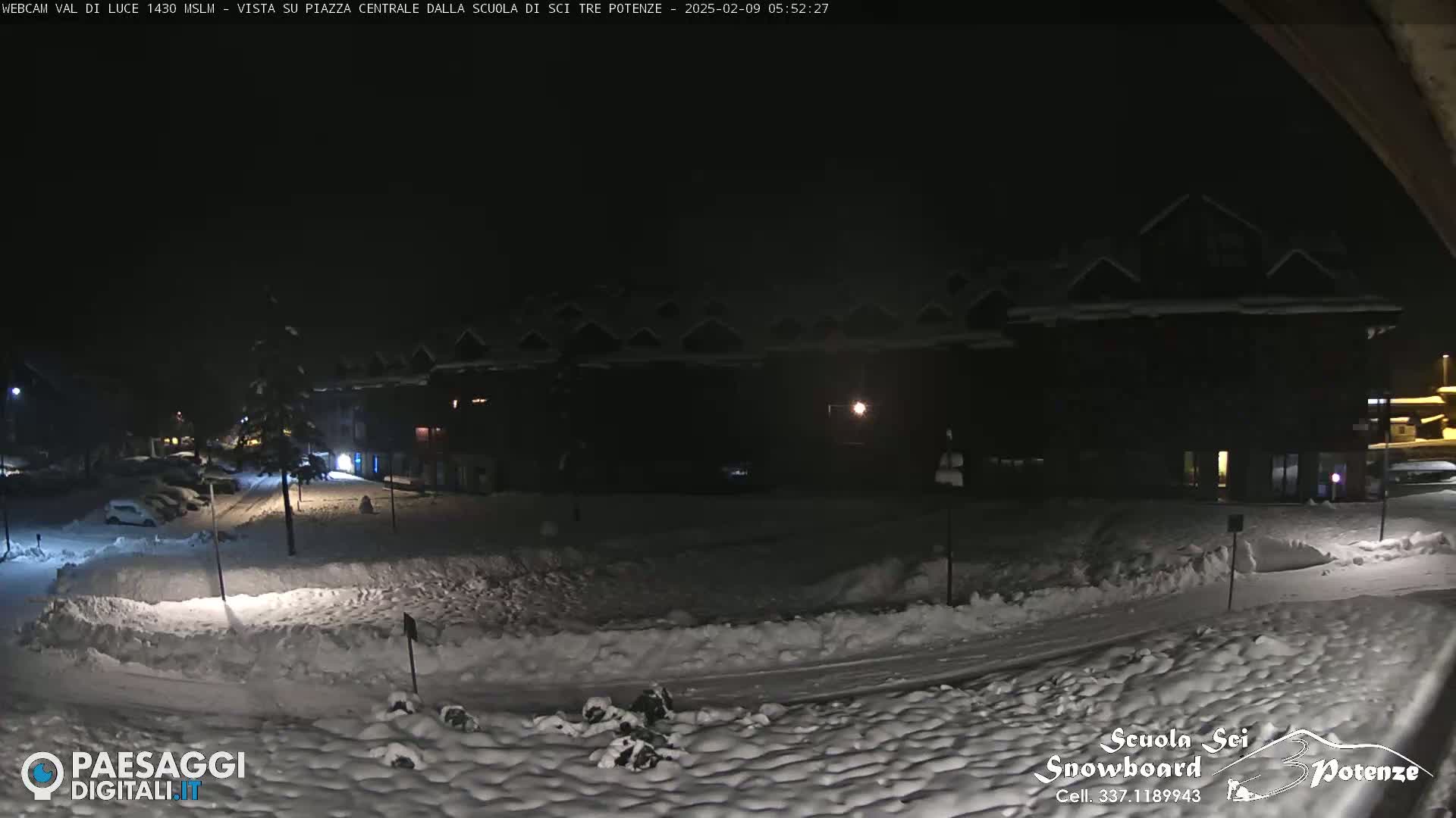 A snow-covered nighttime scene shows a dark building complex and a street with parked cars under a clear, dark sky.