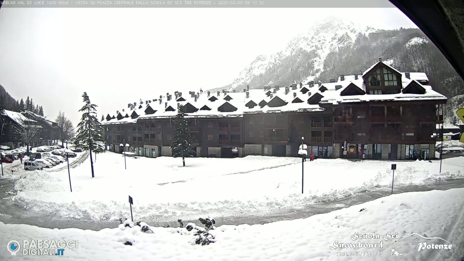 A snow-covered plaza in front of a large, multi-unit building, with mountains in the background under an overcast sky.