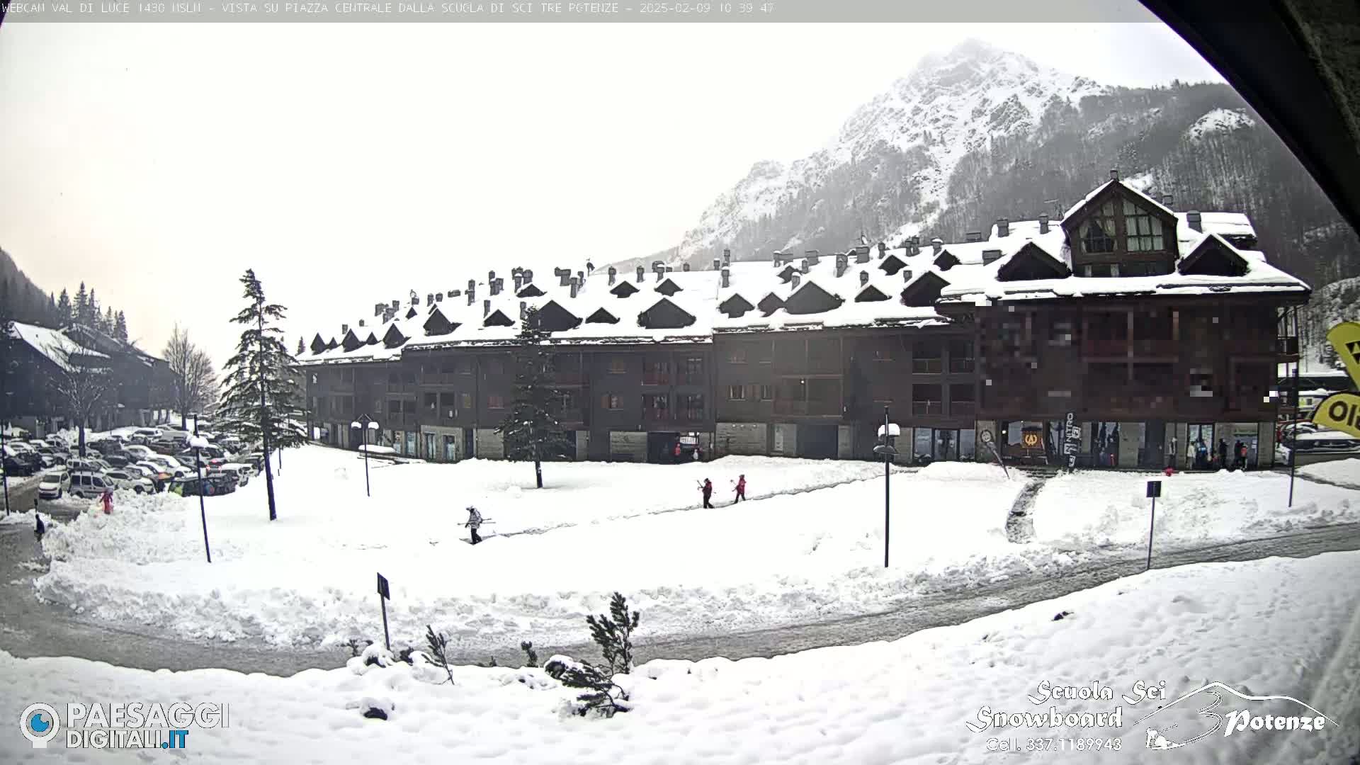 A snow-covered town square with a large, multi-story building, a few pedestrians, and parked cars is seen under an overcast sky in front of snow-capped mountains.