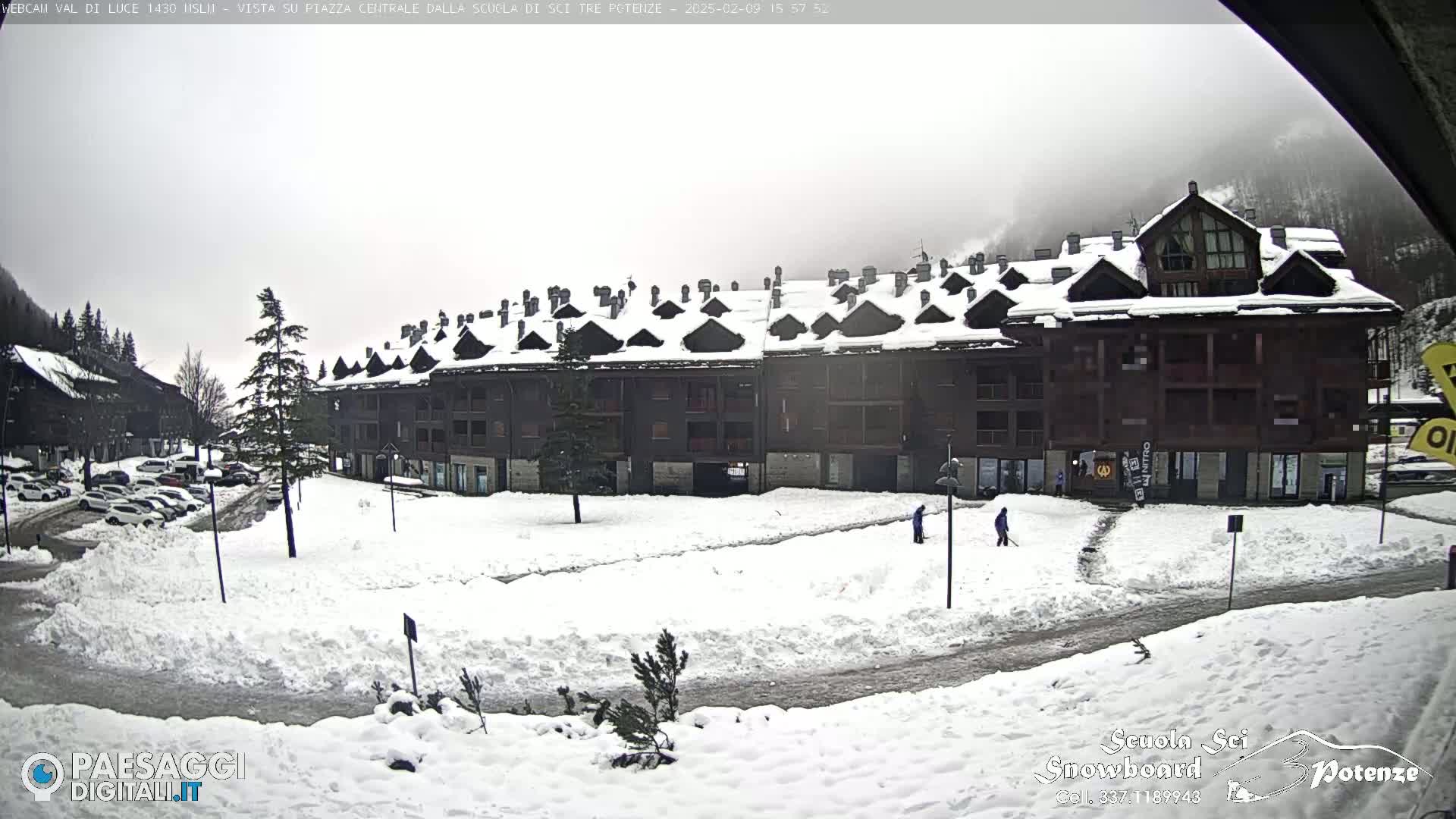 A snow-covered plaza sits in front of a multi-story building, with several people clearing snow from walkways under an overcast sky.