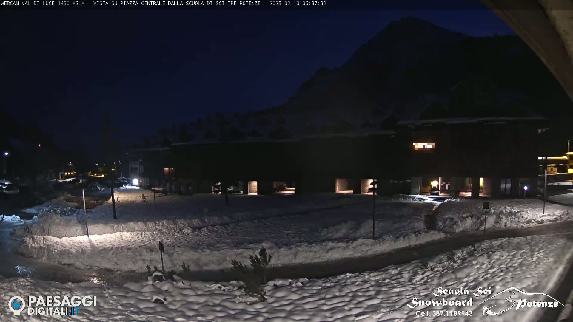 A snow-covered plaza at night, illuminated by artificial lights, is nestled between a dark mountain and a building.