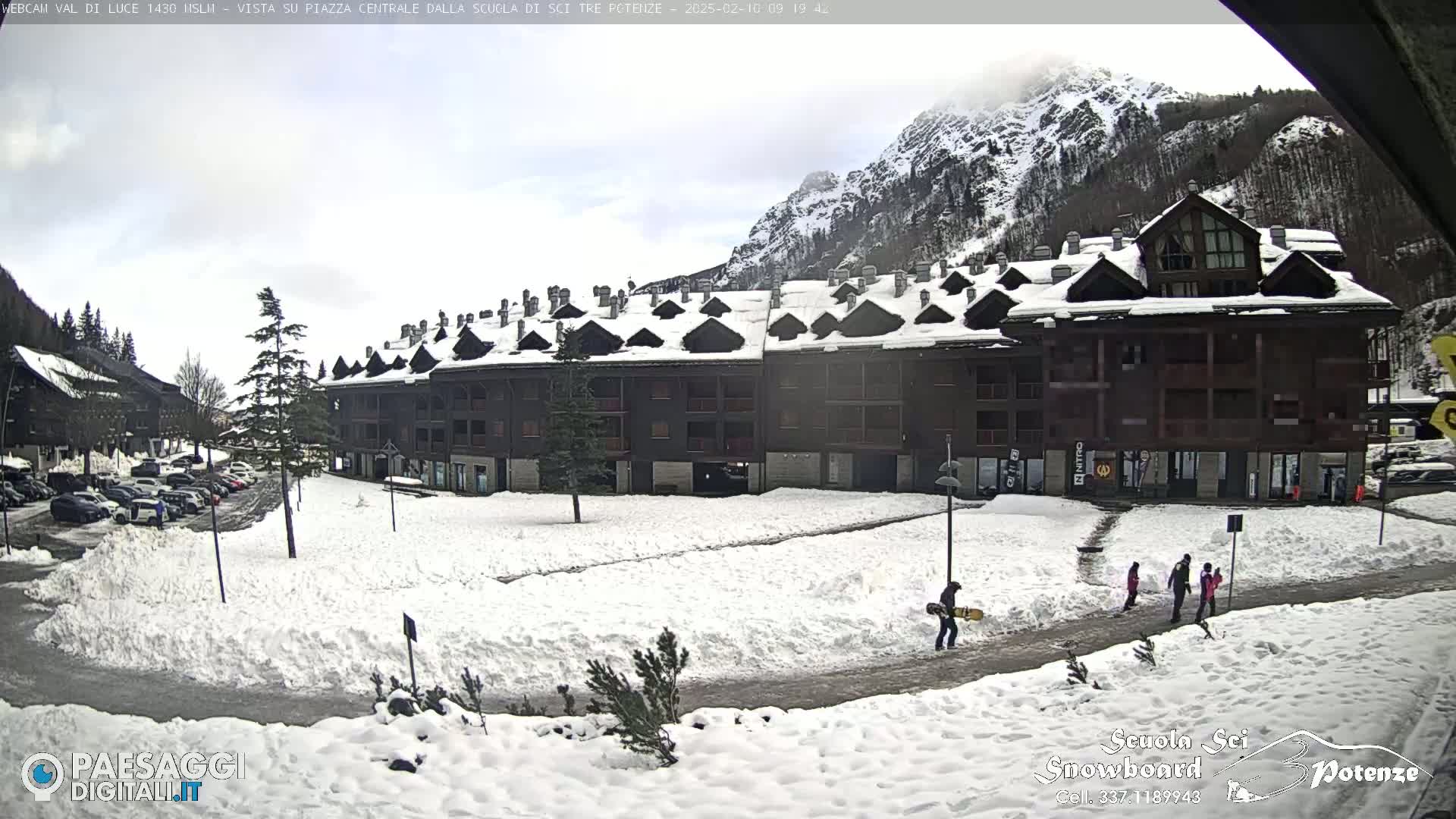 A snow-covered plaza with a multi-story building and several people, including a person carrying a snowboard, is situated in front of a mountain range under an overcast sky.