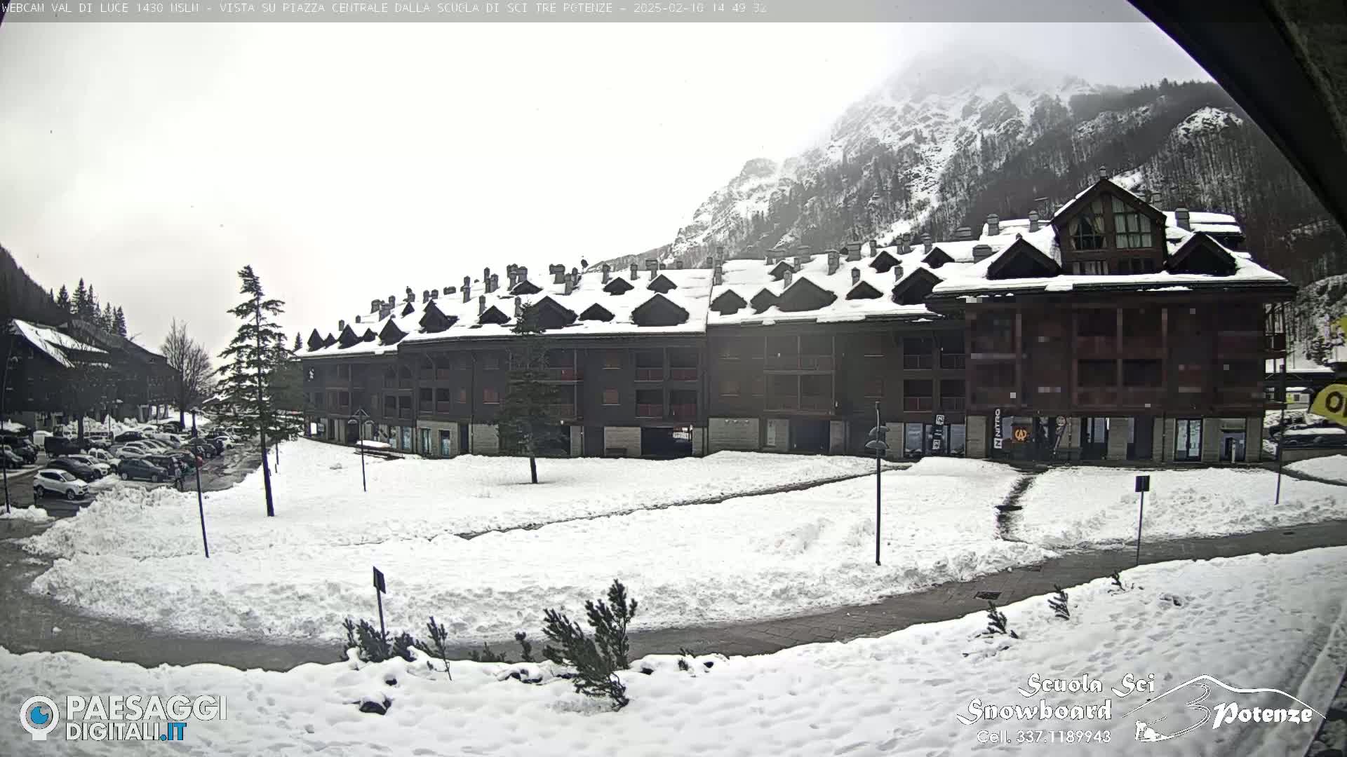 A snow-covered plaza sits in front of a multi-story building, with mountains and overcast skies in the background.